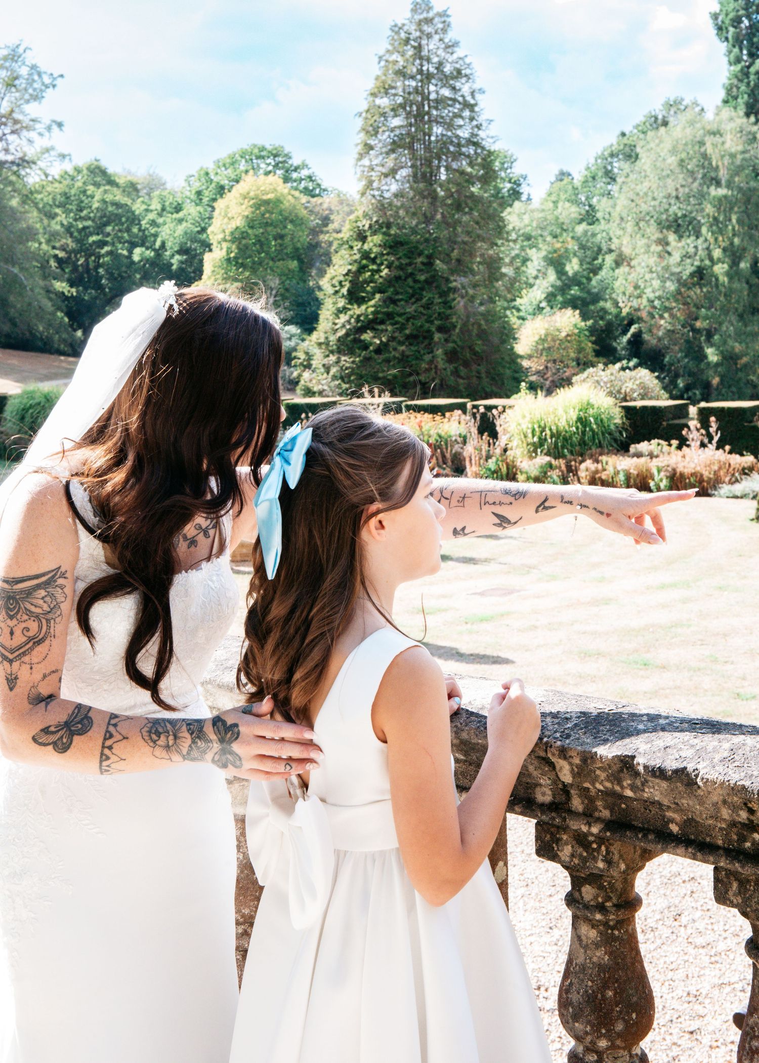 Tattooed bride in white dress and veil stands with flower girl in garden, both looking at lush greenery.