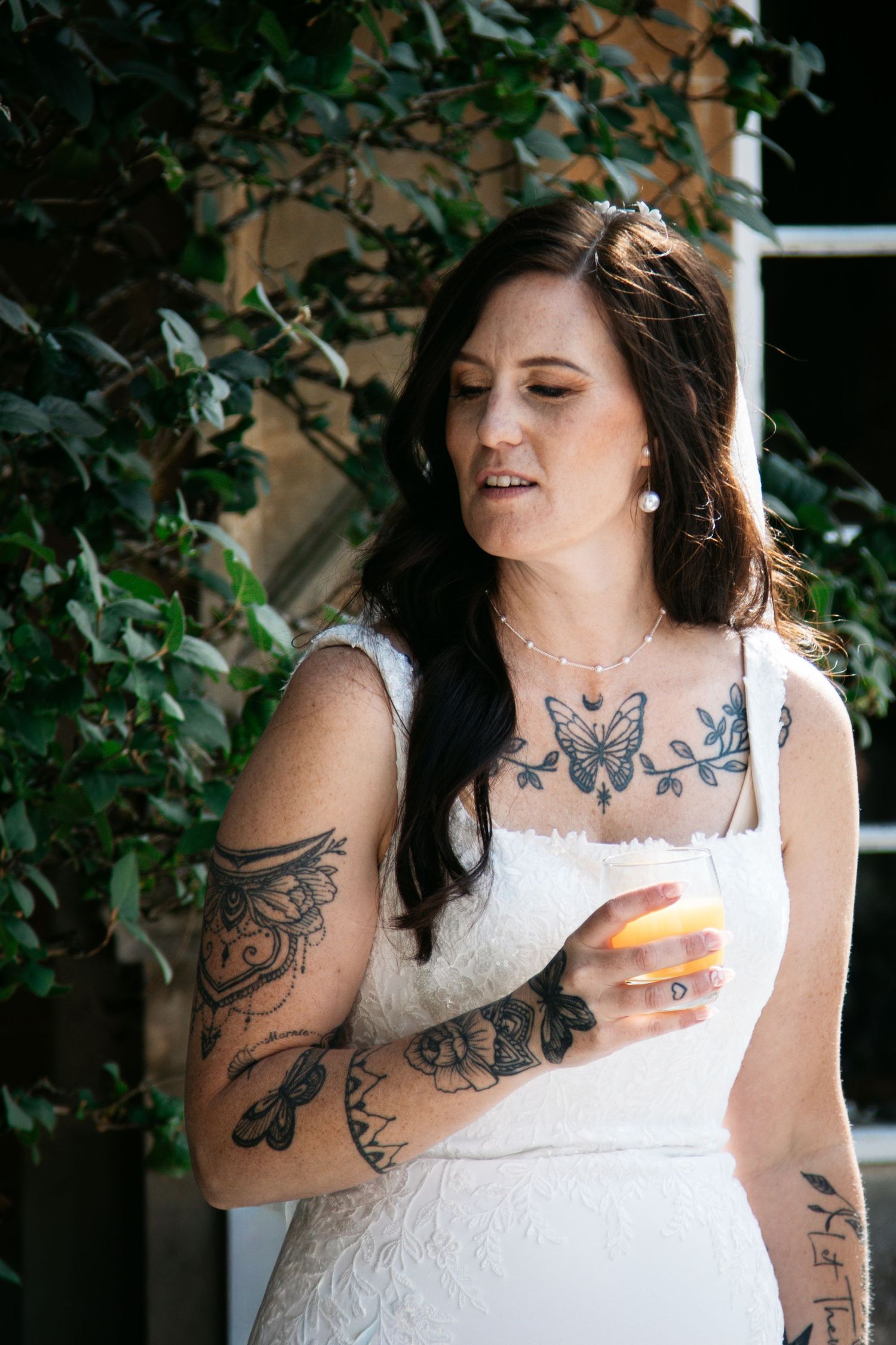 Tattooed bride in white lace dress holding a drink, adorned with floral chest tattoos and pearl jewelry outdoors.