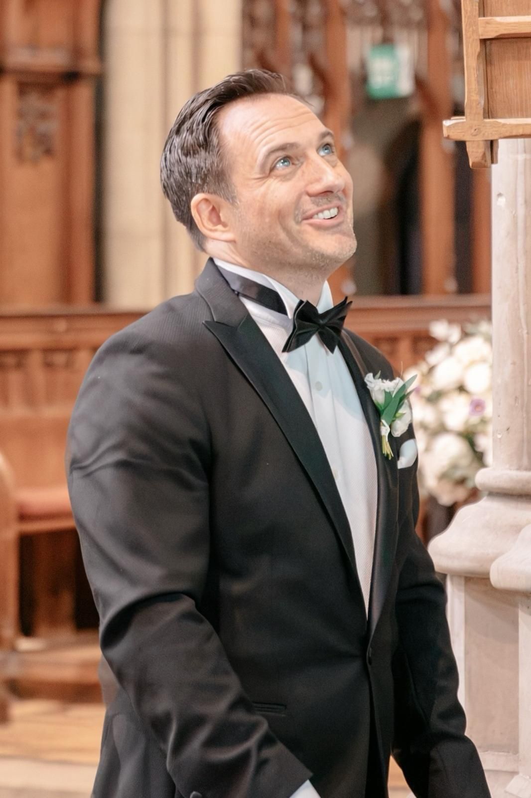 A groom in a black tuxedo with bow tie and boutonniere smiles joyfully inside a church.