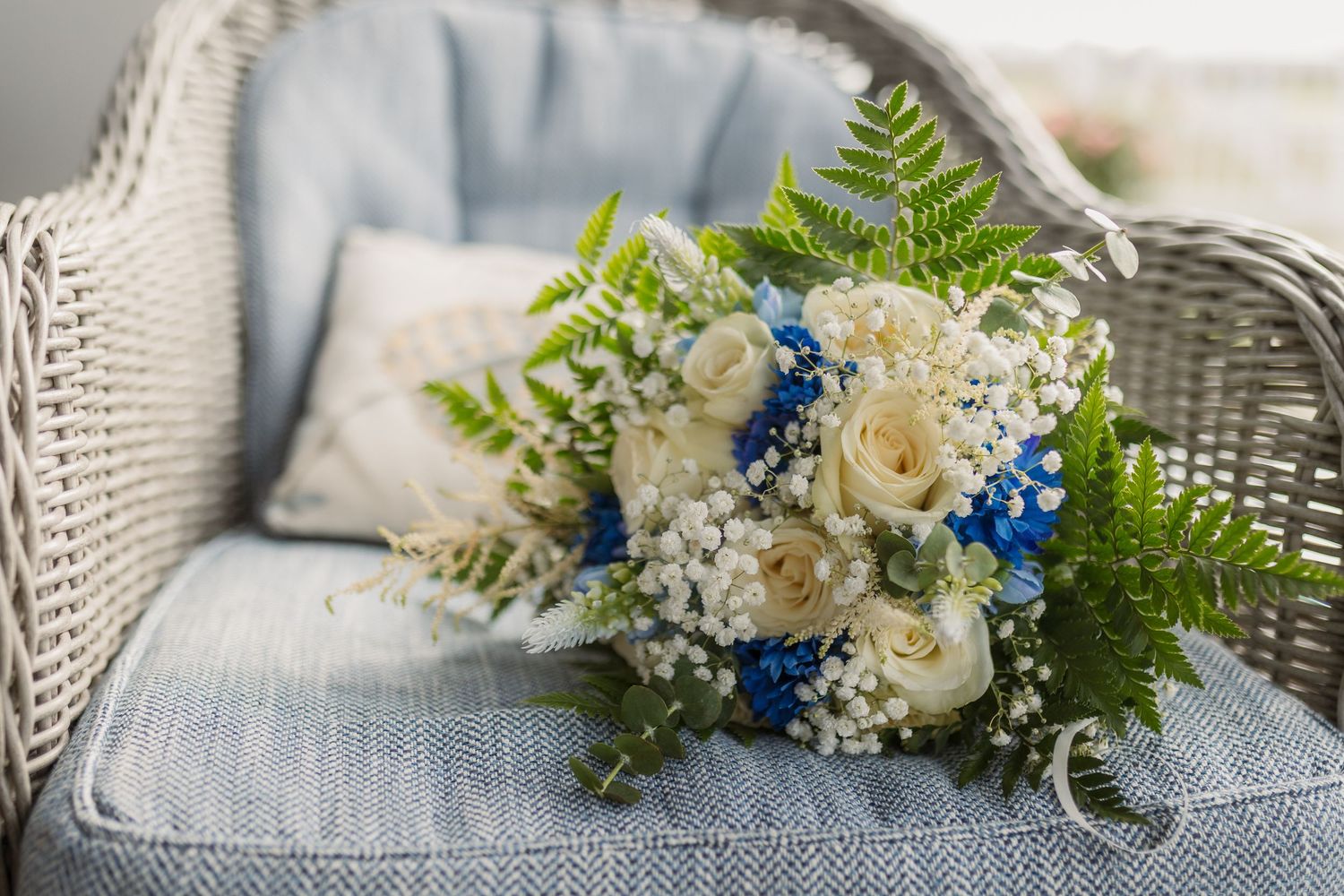 Elegant bridal bouquet with white roses, baby's breath, and blue flowers resting on a wicker chair.