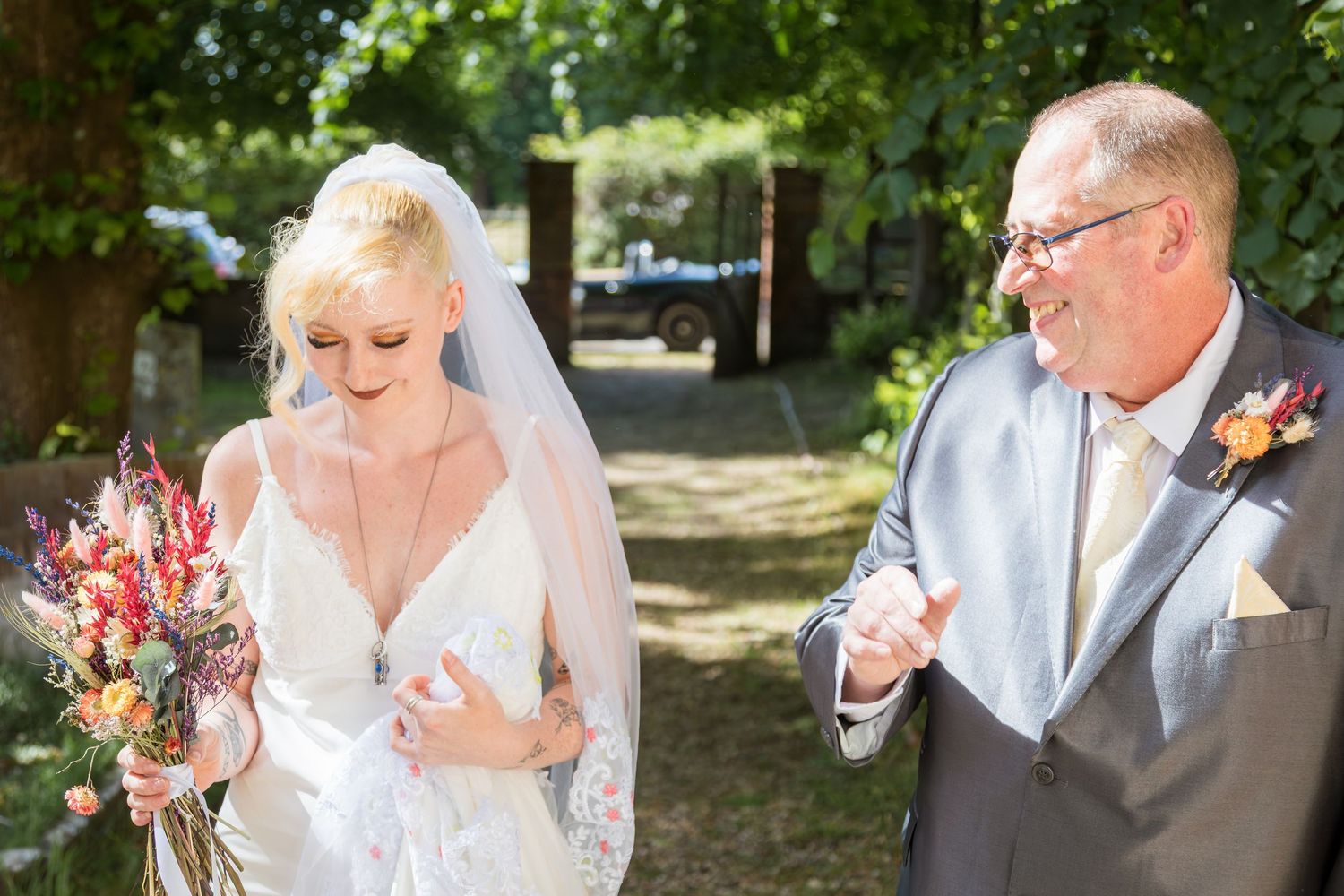 Bride in veil walking with older man holding colorful wedding bouquet outdoors.