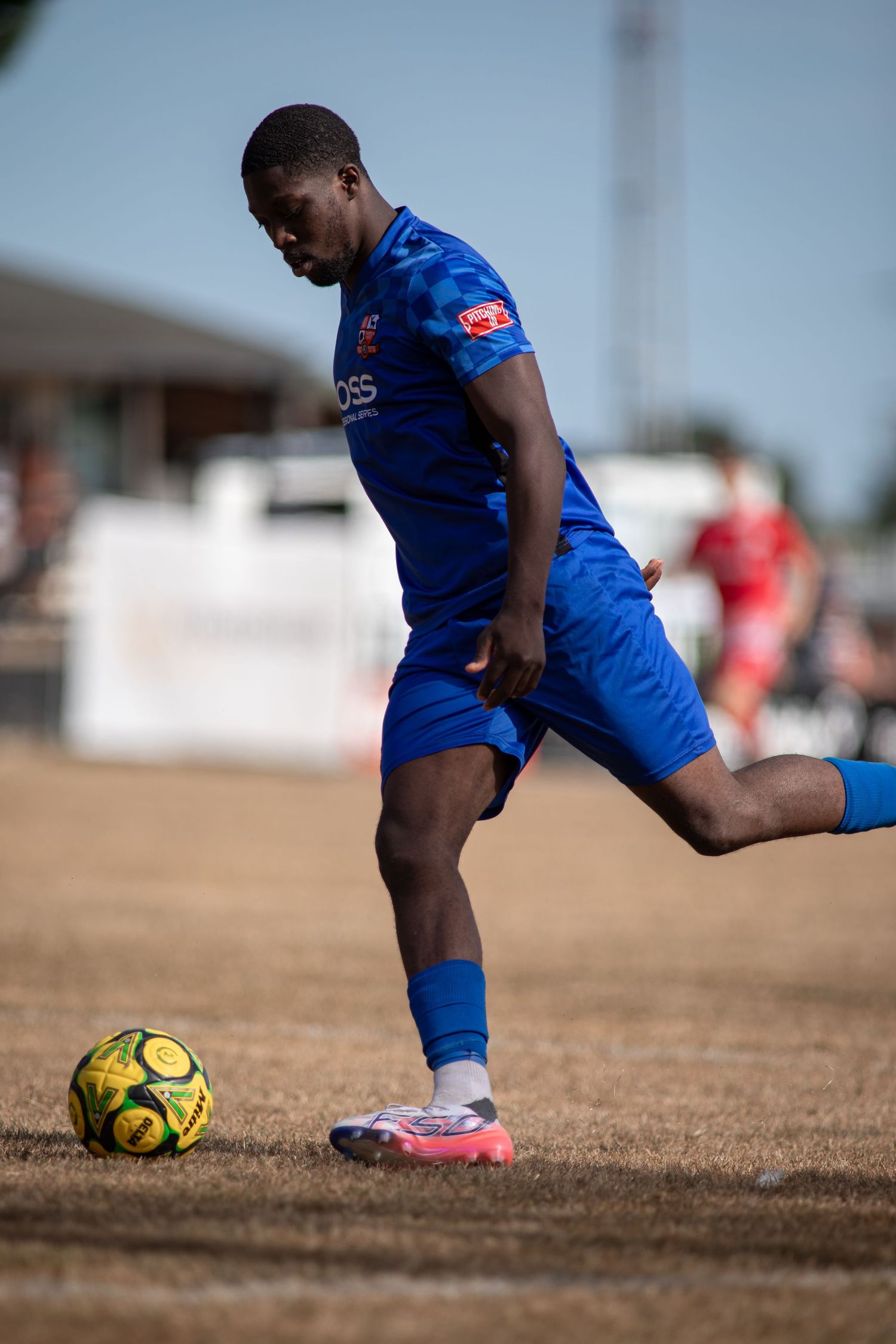 A footballer in a blue BOSS kit prepares to strike a yellow ball on a dry grass pitch.