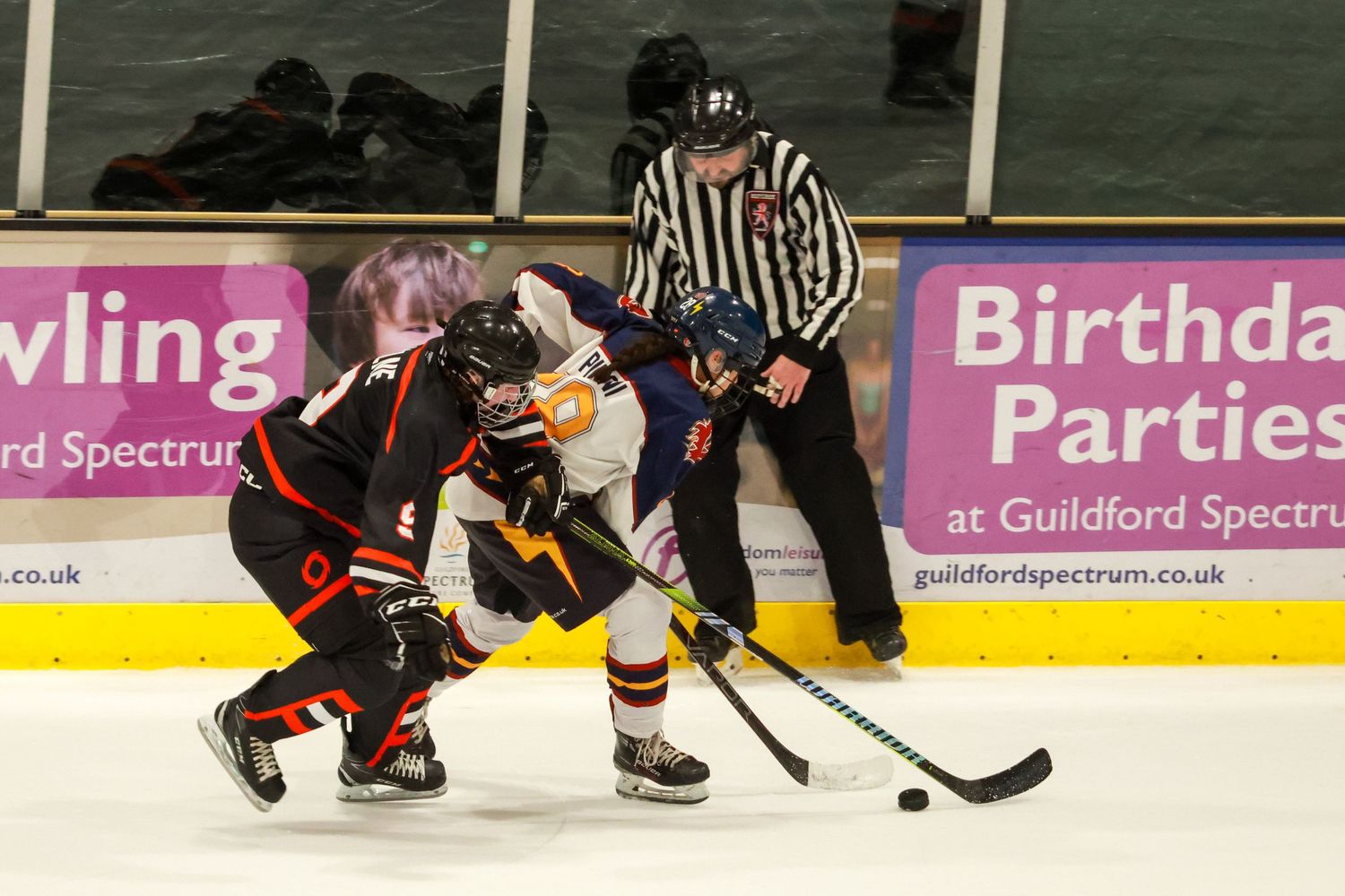 Two ice hockey players face off for puck on ice with referee watching and Guildford Spectrum birthday parties banner behind.