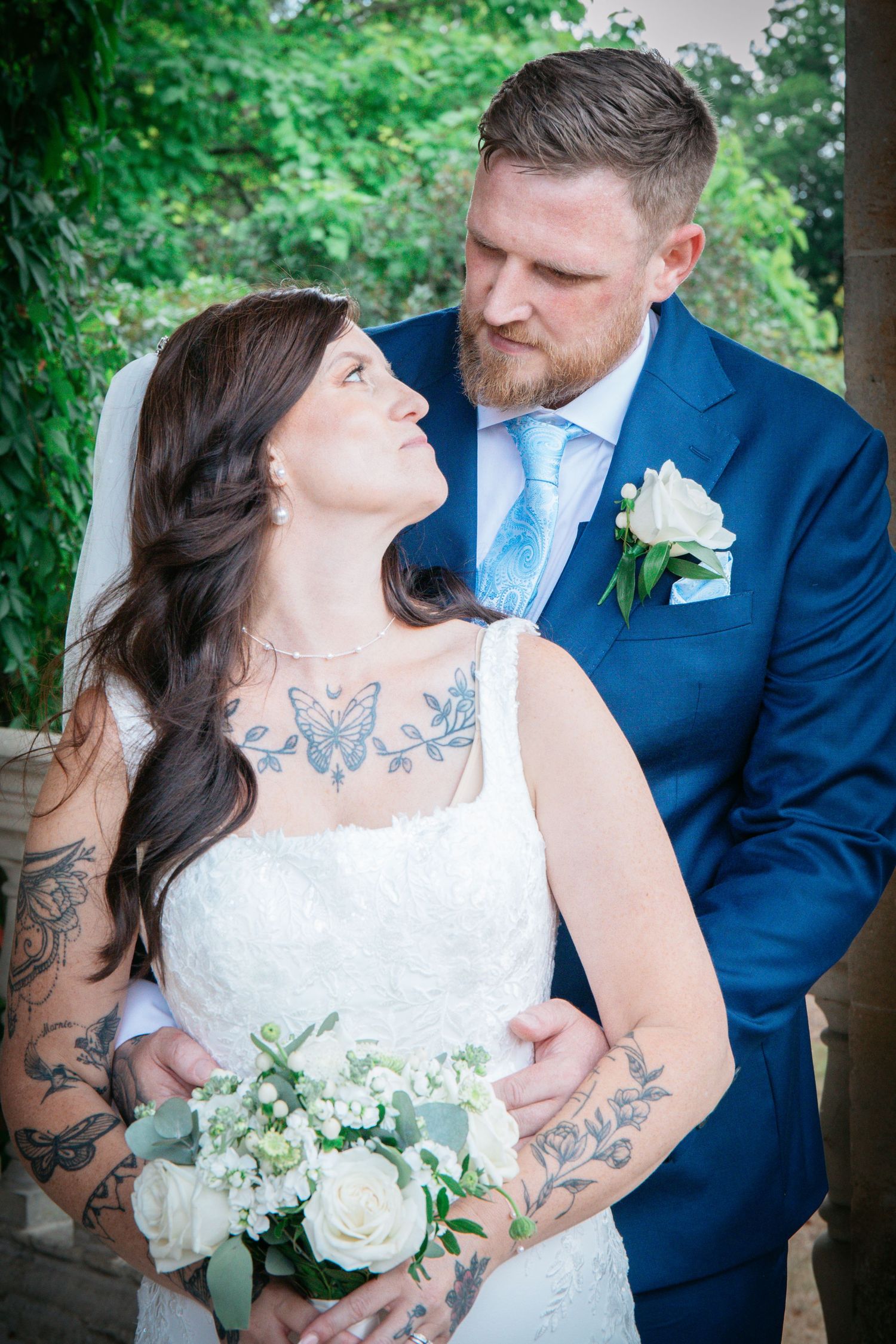 Tattooed bride in white lace gown holds white roses bouquet while groom in blue suit gazes at her outdoors.