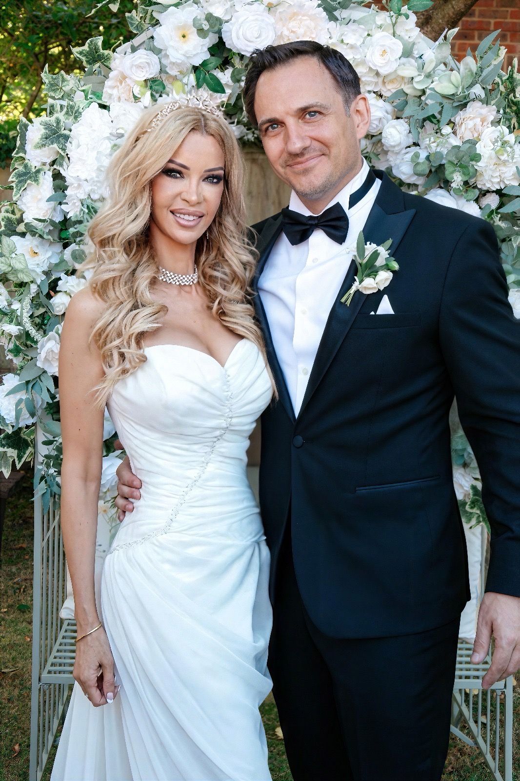 Elegant bride and groom posing before floral arch, bride in white gown with tiara, groom in black tuxedo.