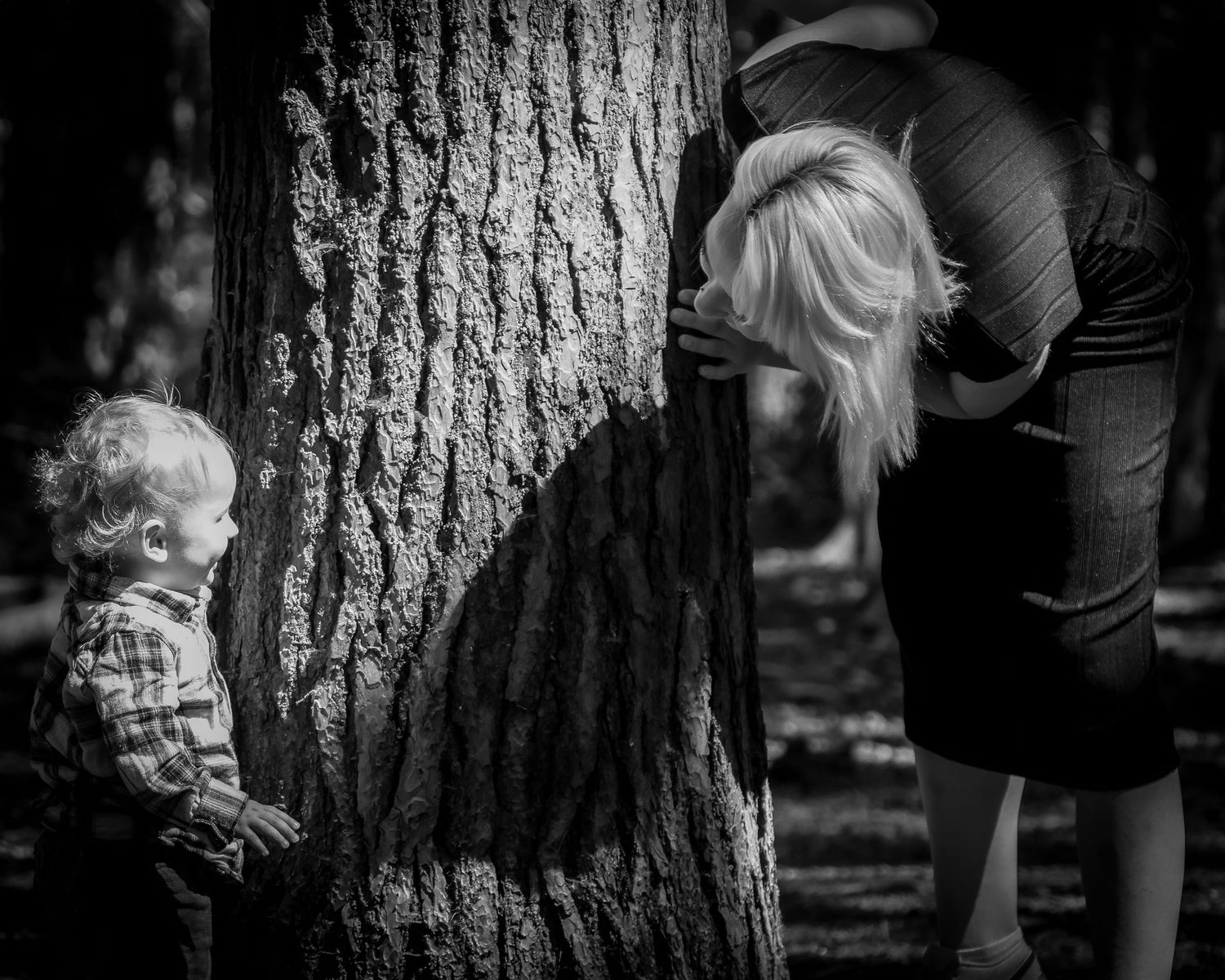 Black and white photo of a toddler boy and his blonde mother peeking around a large tree.