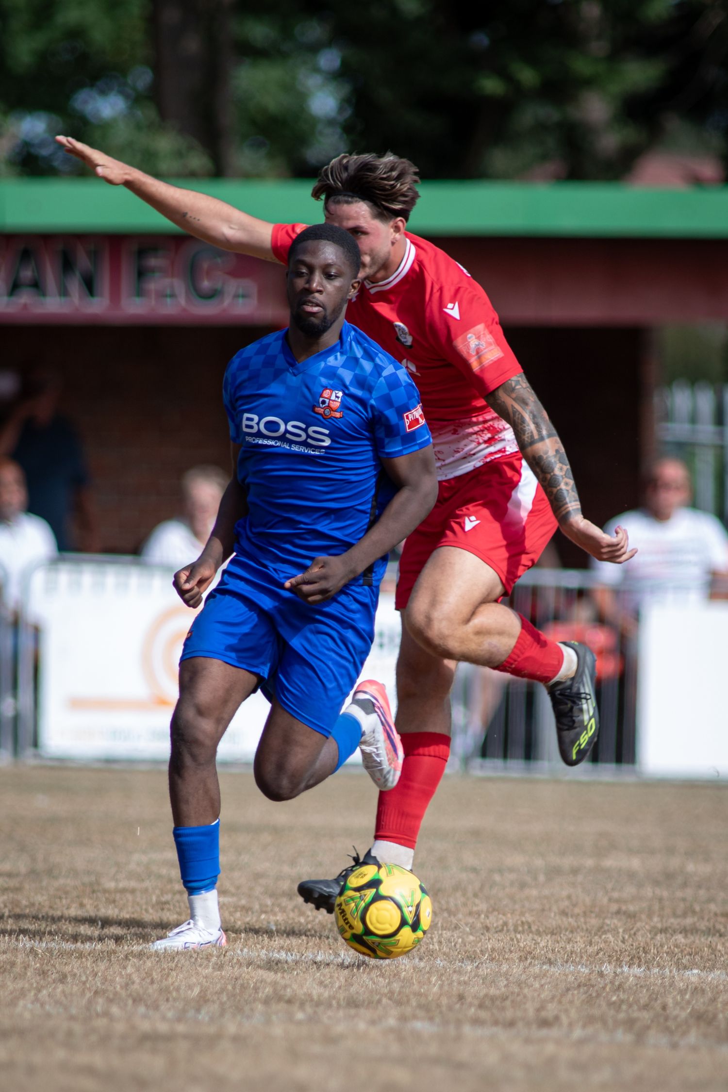 Two football players battle for possession, one in blue BOSS kit and one in red, during a match.
