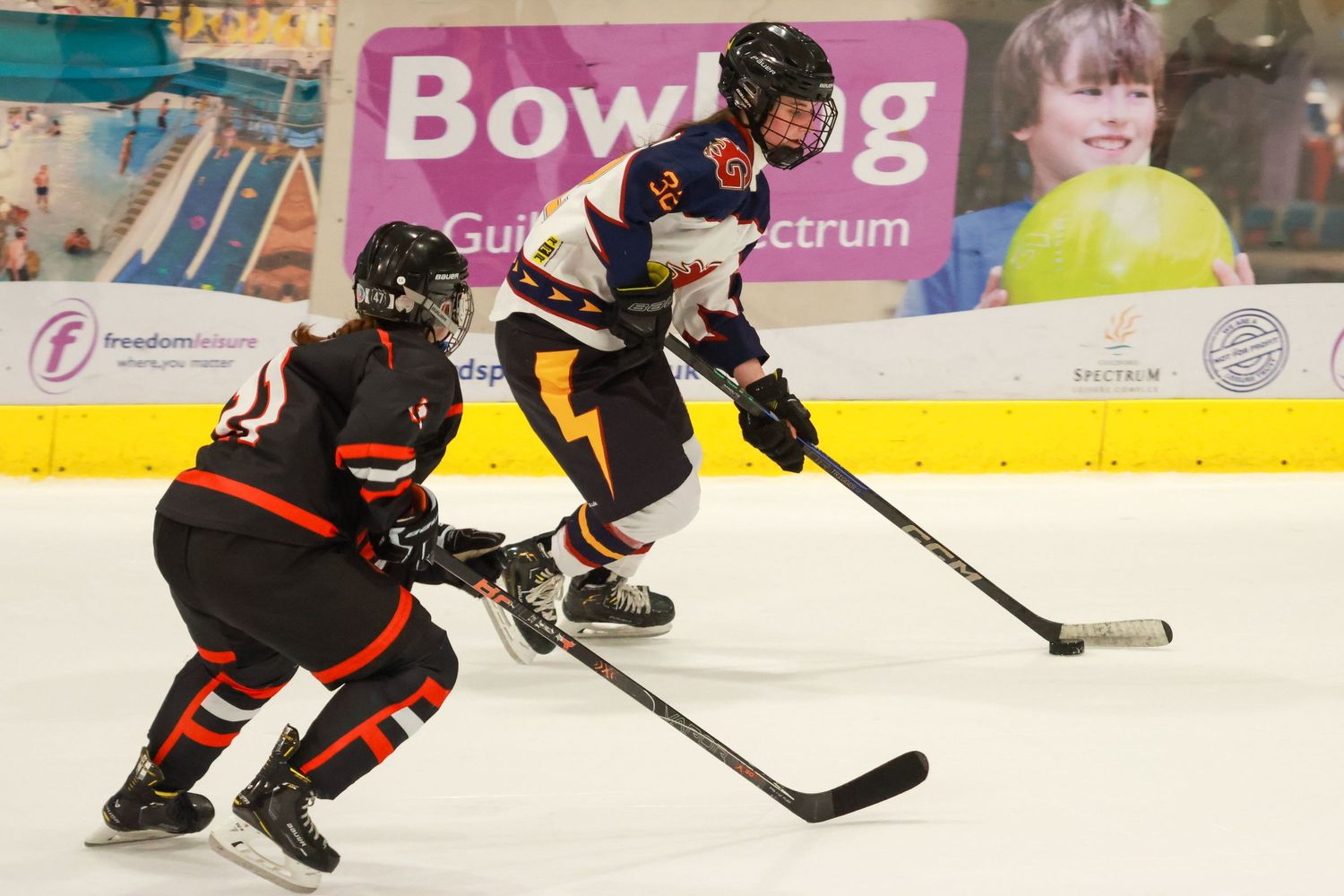 Female ice hockey player in white uniform controls puck while defender in black pursues near Guildford Spectrum bowling banner.