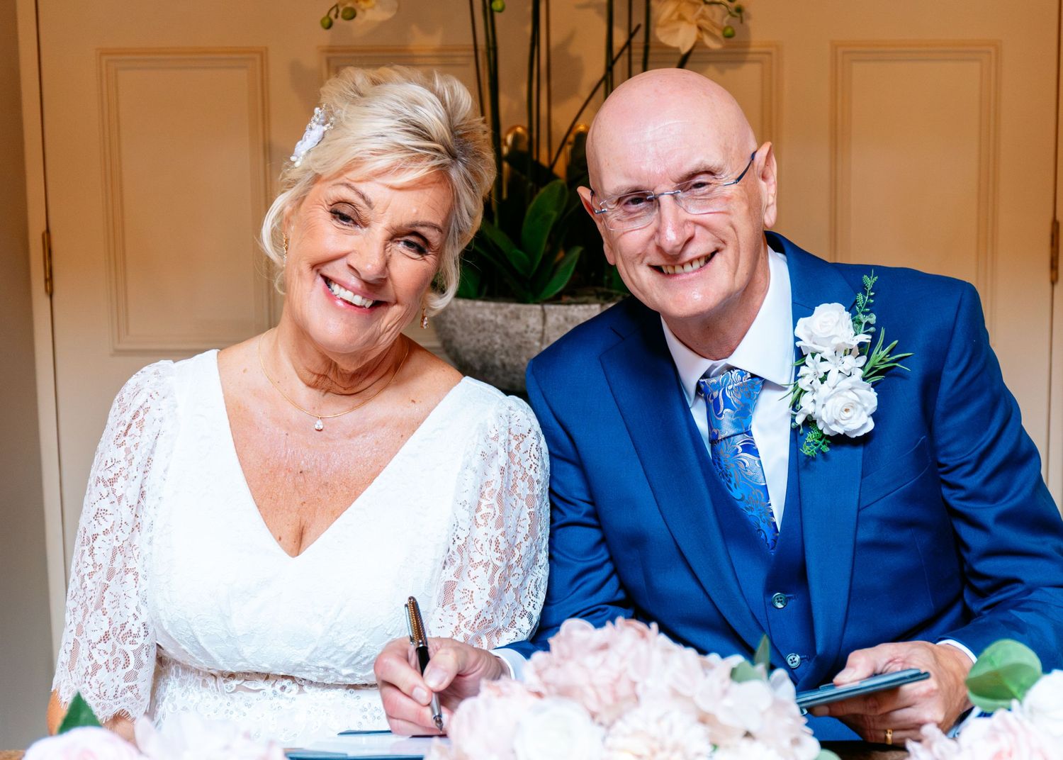Happy couple signing wedding register, bride in white lace dress and groom in blue suit with white boutonniere.