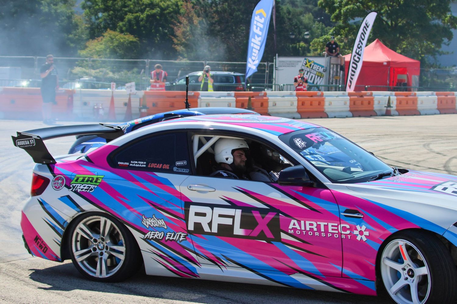 Pink and blue RFLX Airtec sponsored BMW drift car powerslides at a motorsport competition.