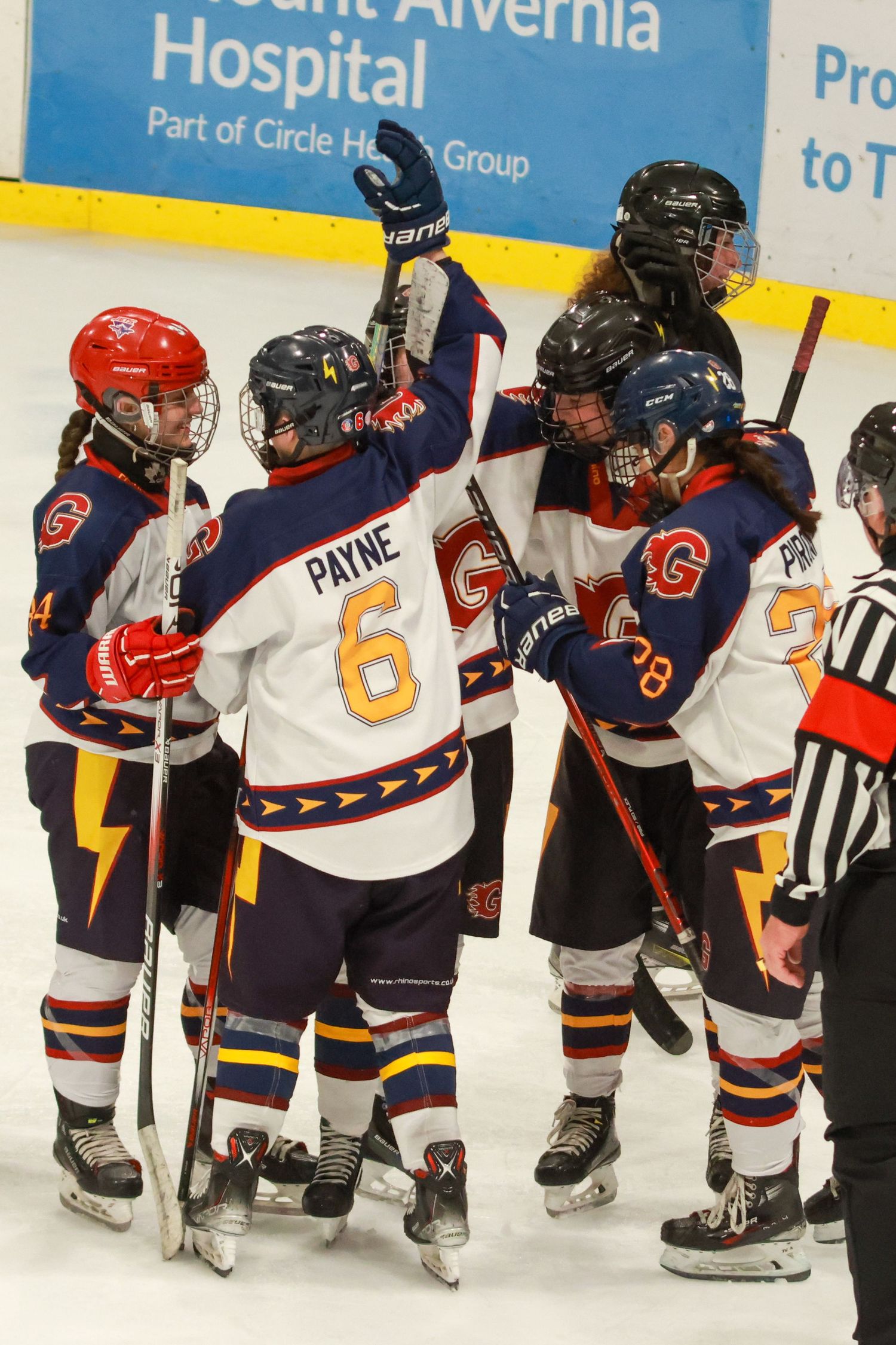 Female ice hockey players celebrate on the rink, with player Payne wearing number 6 jersey, near Mount Alvernia Hospital ad.