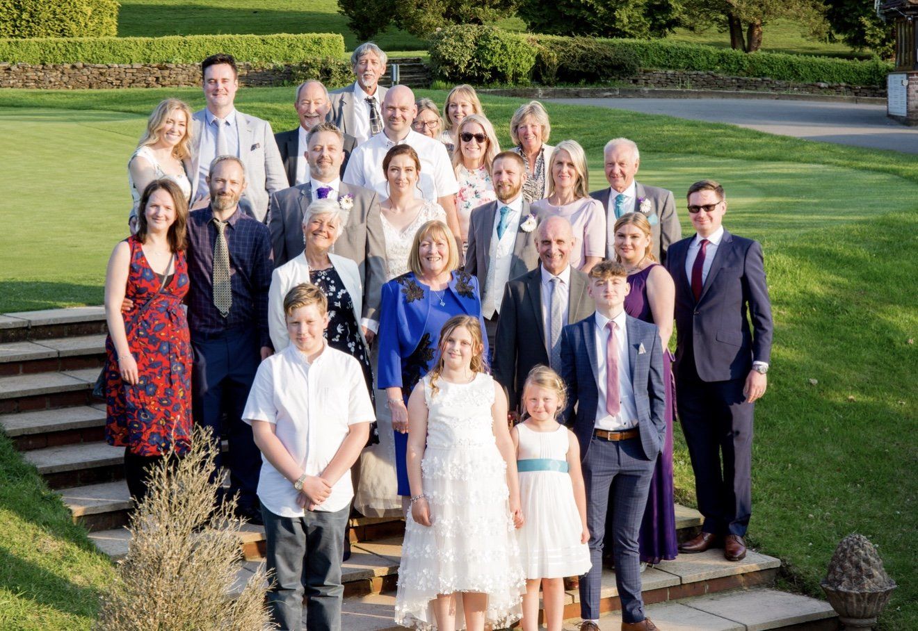 Large wedding family group portrait posed on garden steps in warm evening sunlight outdoors.