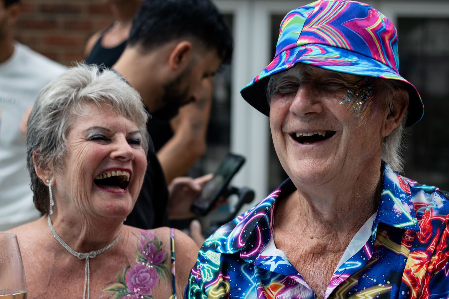 An older couple laughs joyfully together outdoors, the man wearing a colorful bucket hat and tropical shirt.