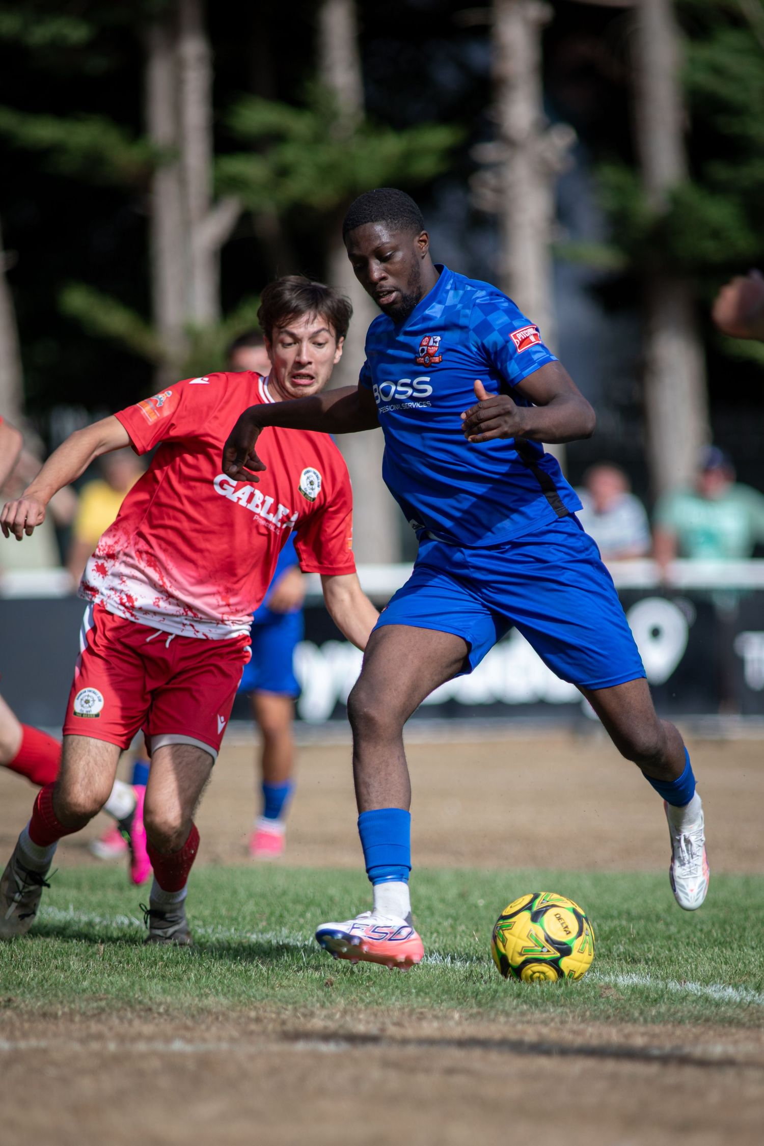 Blue BOSS jersey footballer shields ball from red-kit opponent during competitive soccer match.