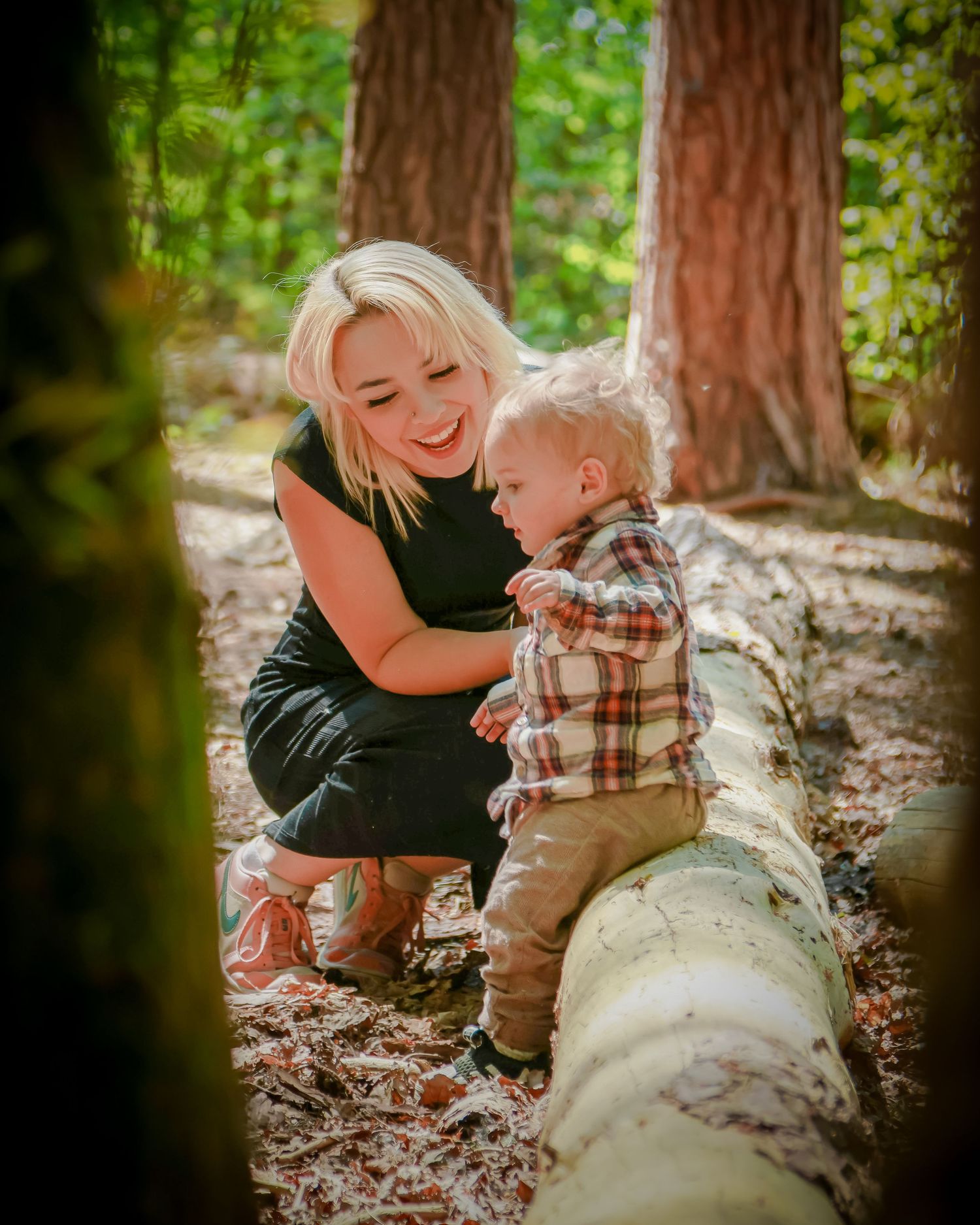 Blonde mother smiling at her toddler son near a fallen log in a bright sunlit forest.