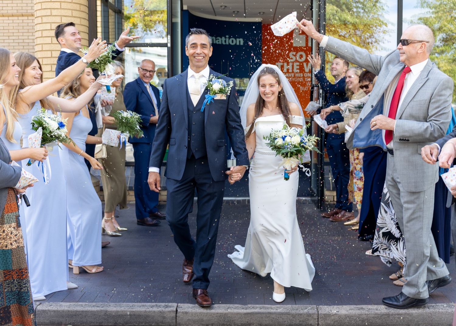 Newlywed couple exits wedding venue holding hands while guests celebrate with confetti in sunny outdoor ceremony.