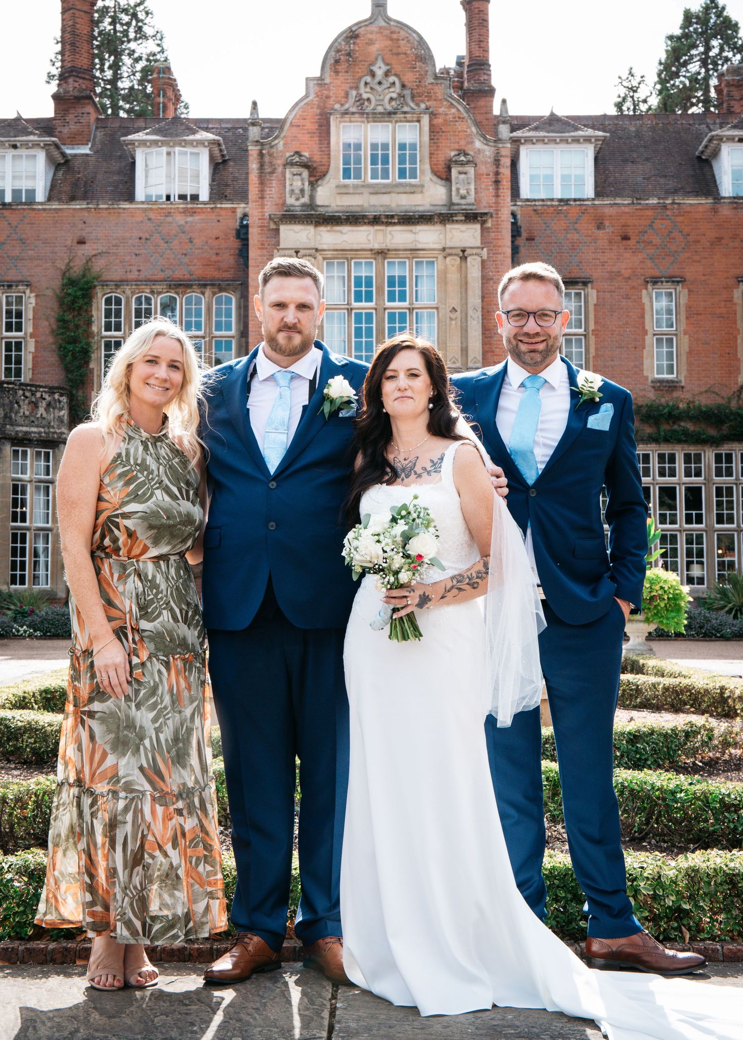 Wedding party of four posing outside a grand red brick manor house on a sunny day, bride holding white bouquet.