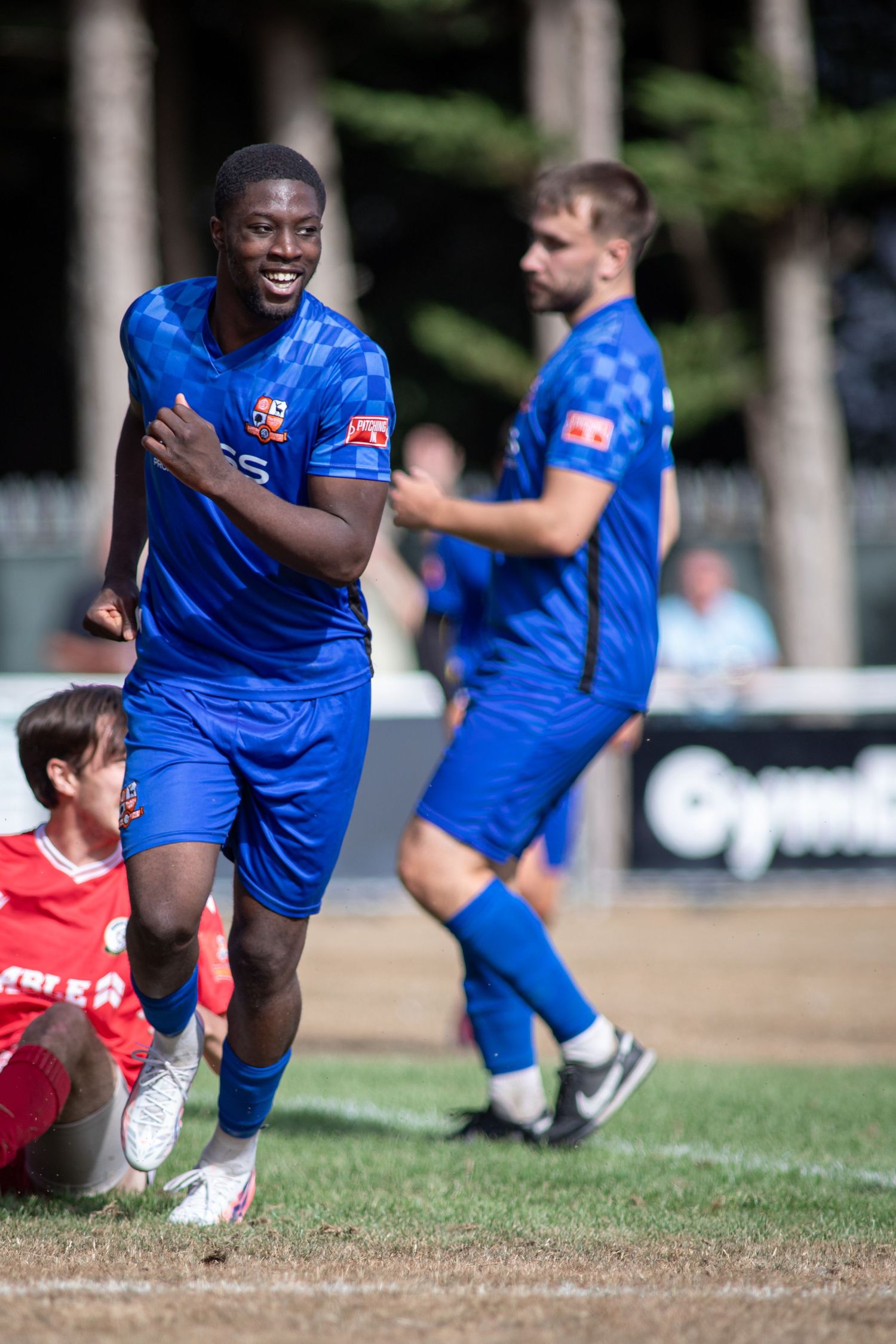 Smiling blue-kit footballer celebrates near fallen opponent with teammate approaching in background.