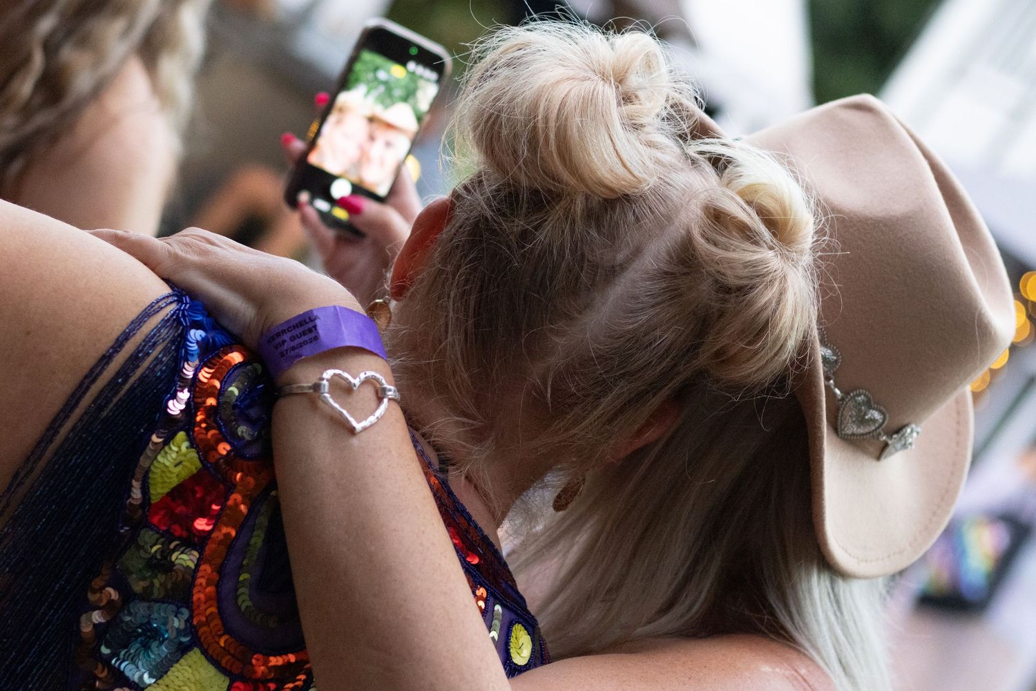Blonde woman wearing cowboy hat and heart arm tattoo using smartphone at outdoor festival.