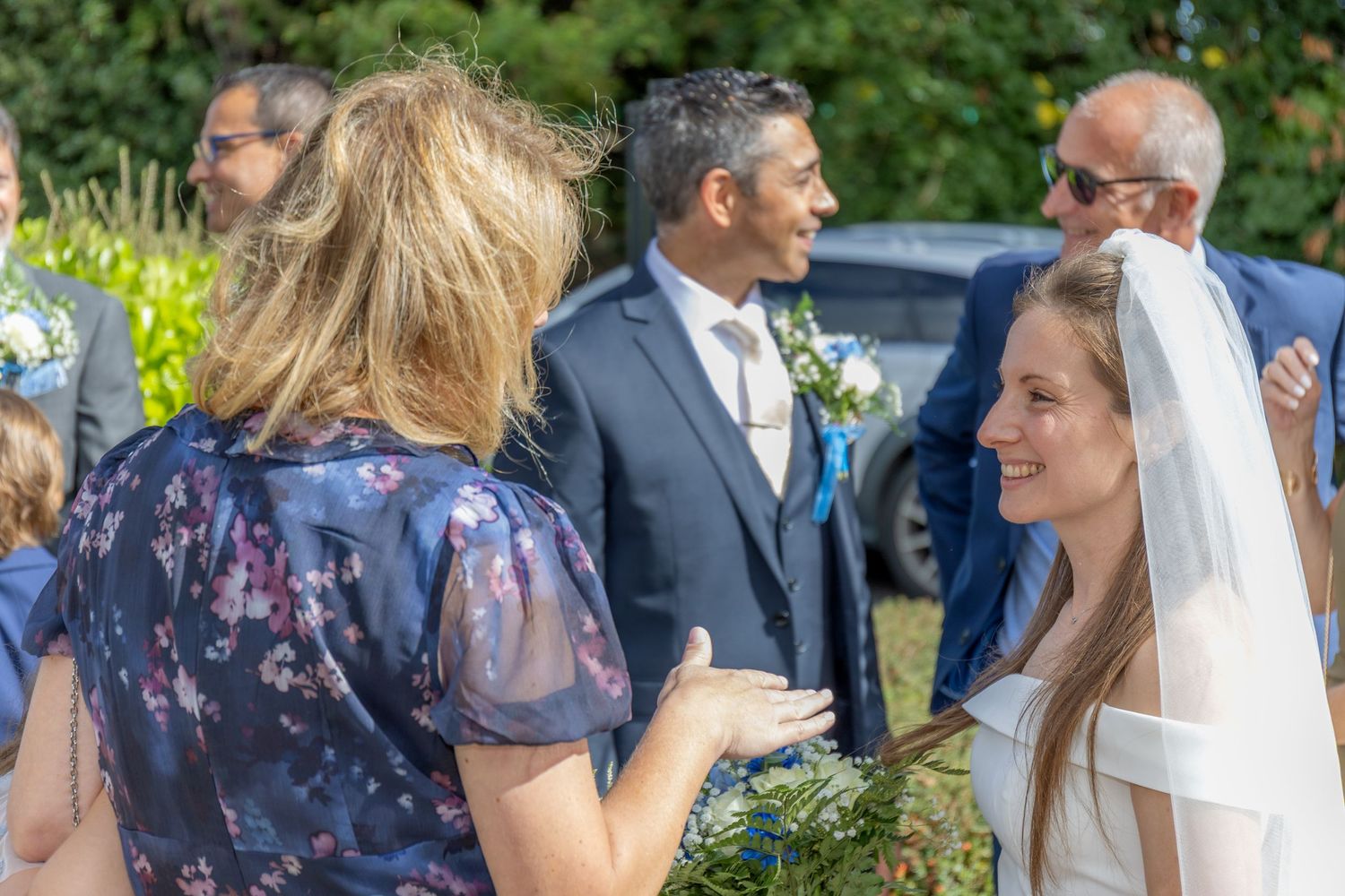 Bride in white veil smiling with guests at an outdoor wedding celebration on a sunny day.