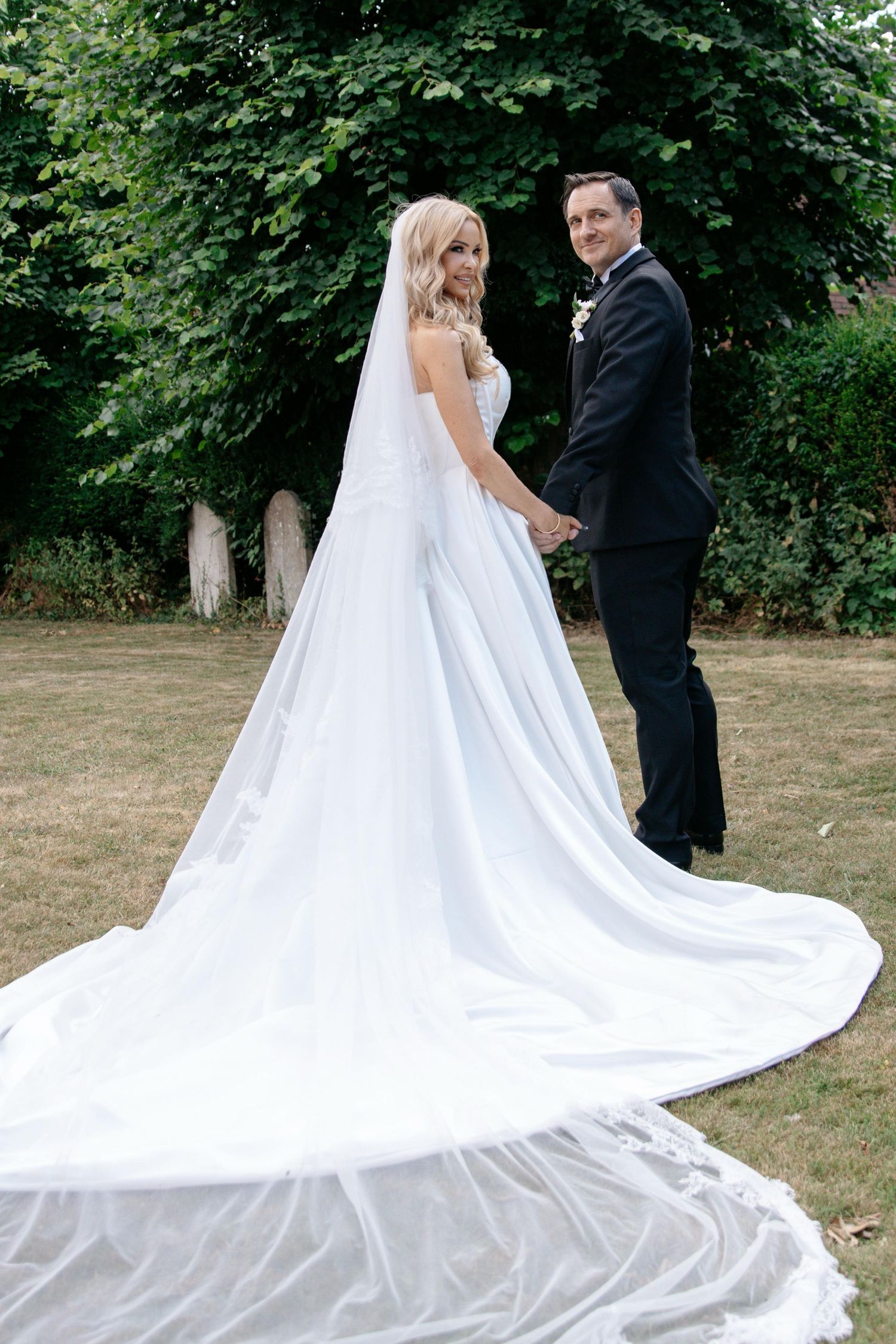 Bride in long white gown with cathedral veil holds hands with groom in black suit outdoors.