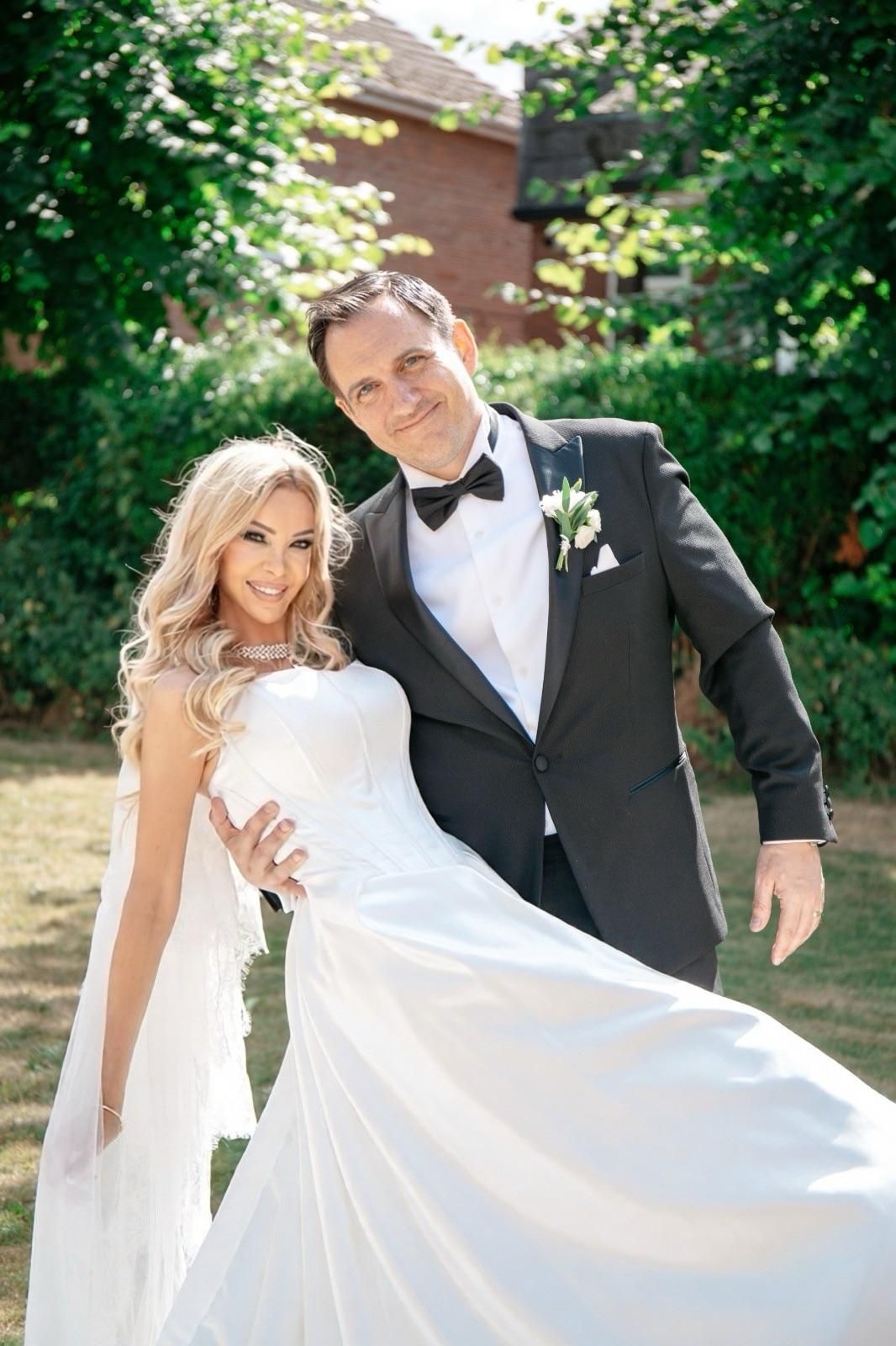 Bride in white gown and groom in black tuxedo posing together outdoors on their wedding day.