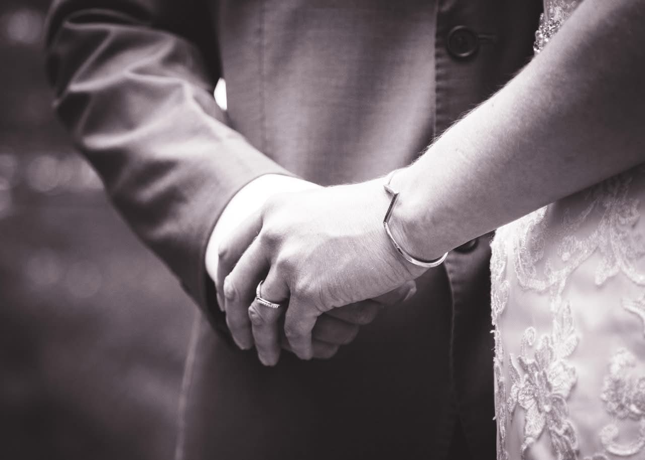 Black and white photo of a bride and groom holding hands, showing wedding rings and bridal lace dress.