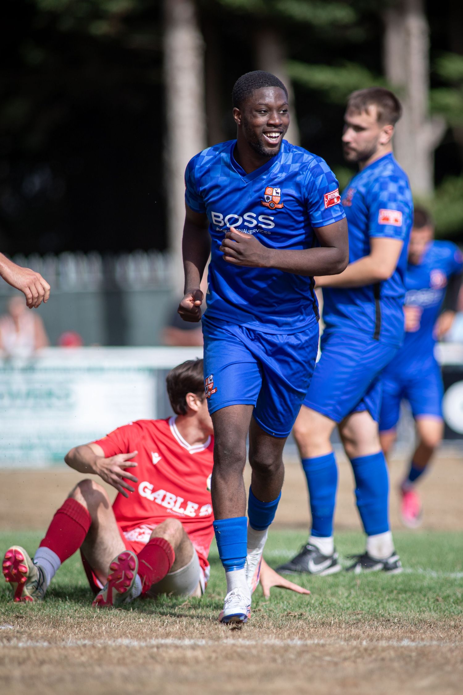 Blue BOSS jersey footballer celebrates near fallen red-kit opponent during outdoor soccer game.