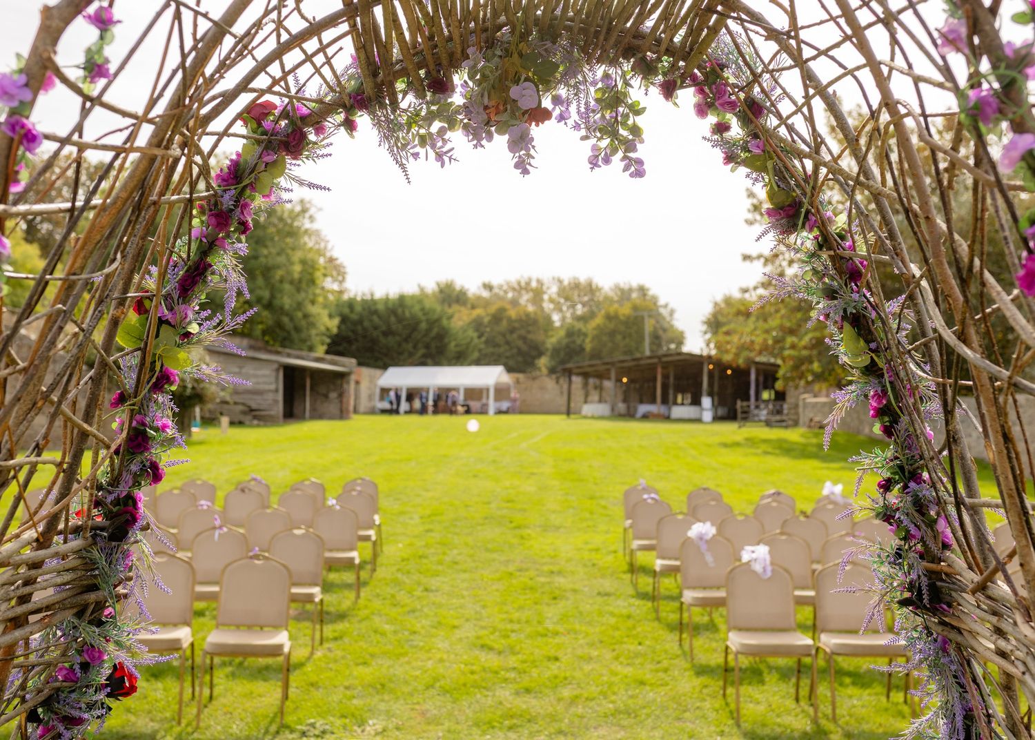 Rustic wicker arch adorned with purple flowers frames outdoor wedding ceremony chairs on green lawn.