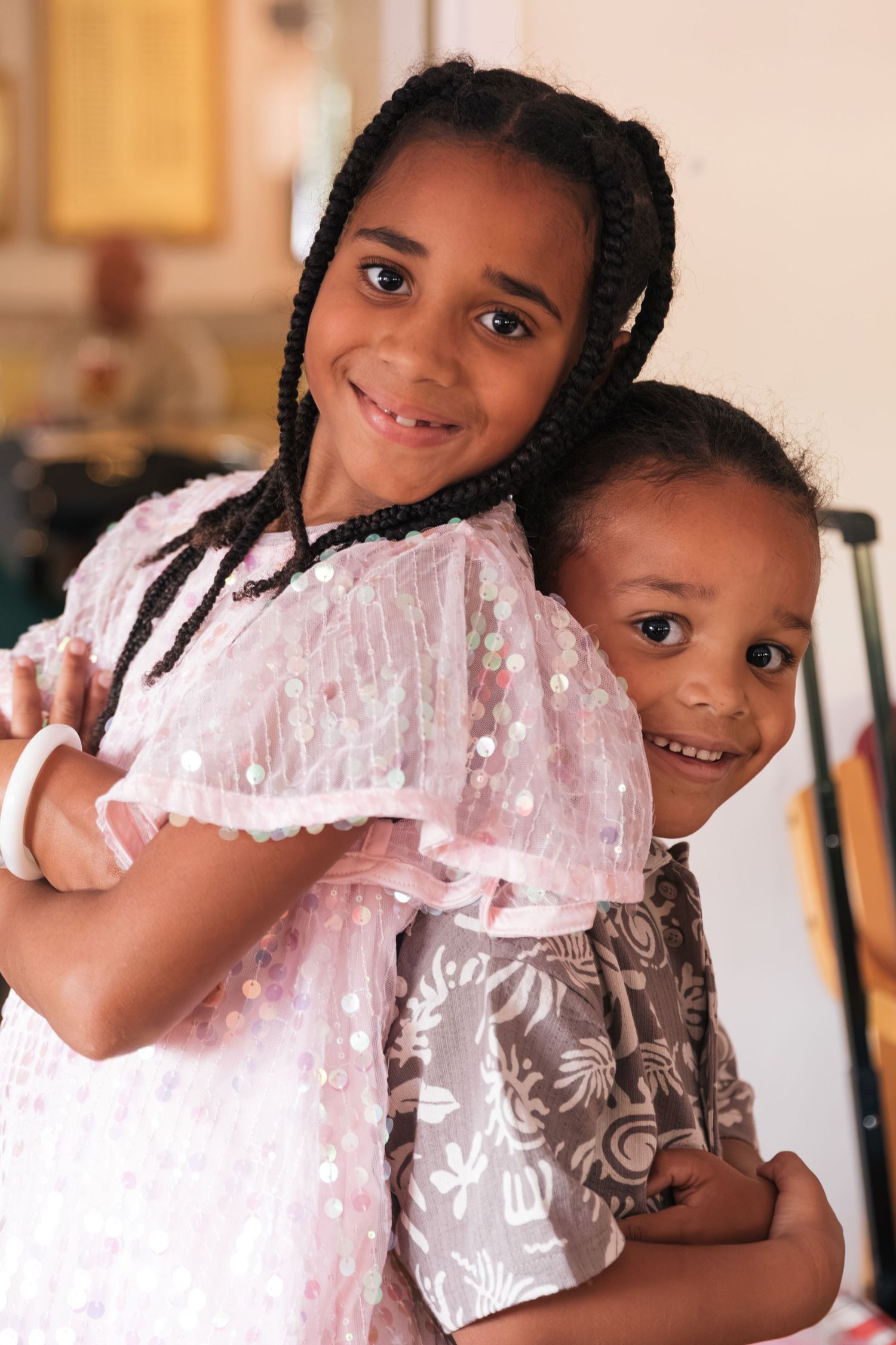 Two smiling African American children hugging together, girl in pink sparkly dress with braids.