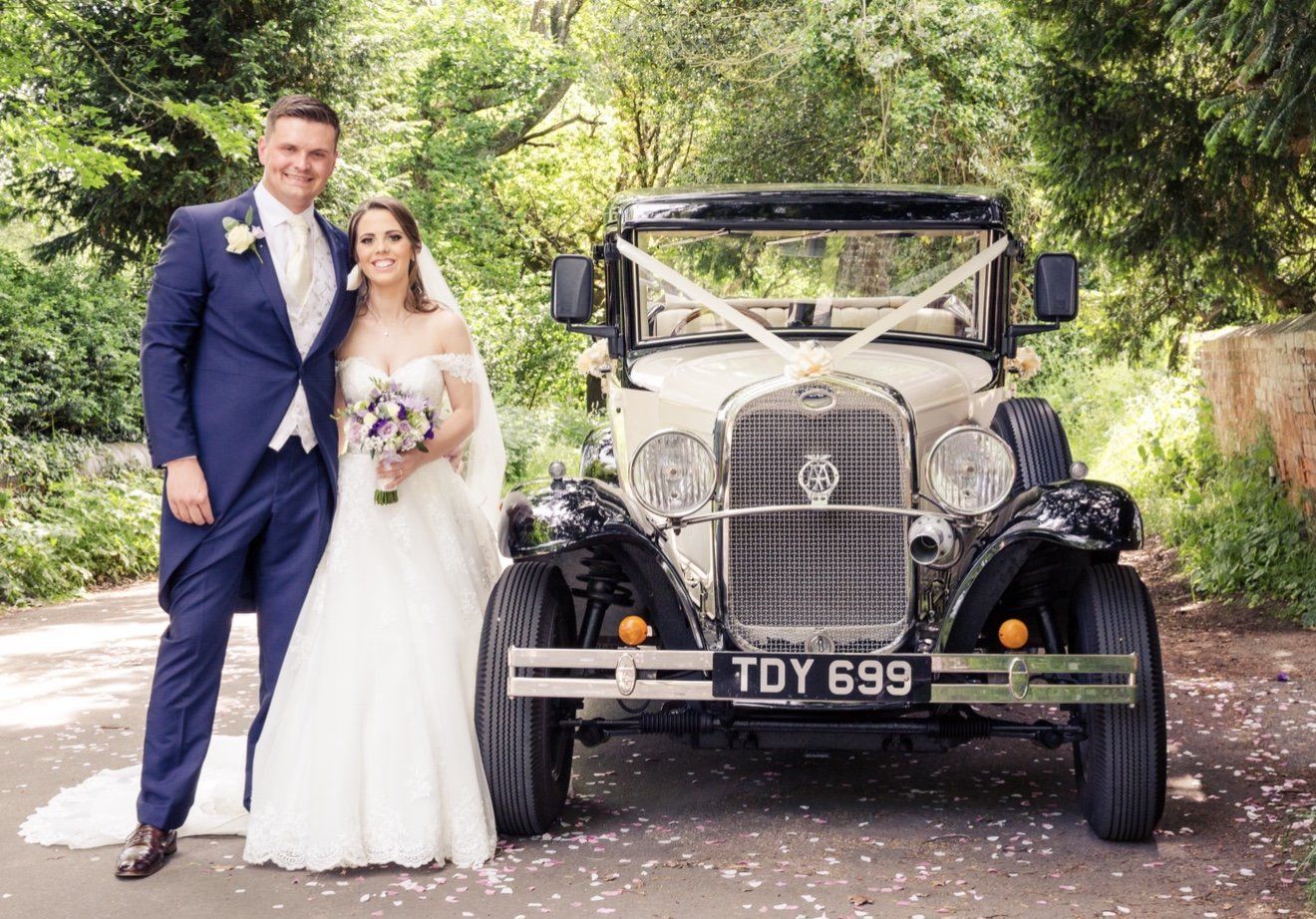Bride and groom posing beside a vintage classic wedding car decorated with ivory ribbons outdoors.