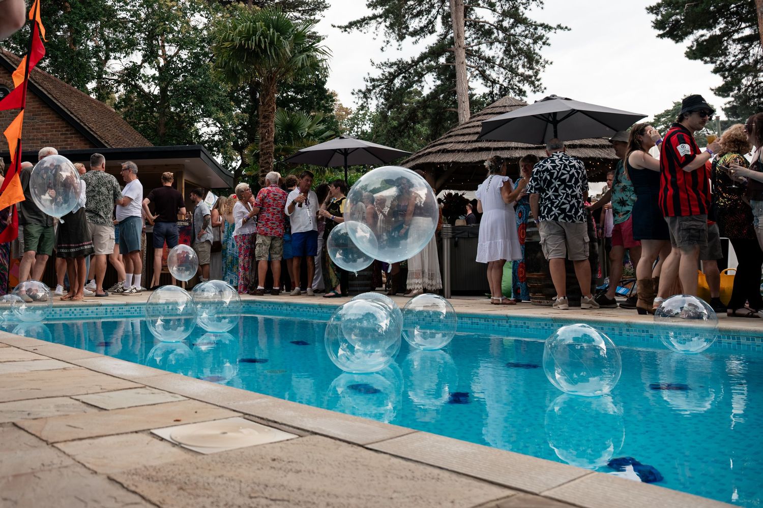 Large transparent balloons float over a swimming pool at an outdoor garden party with guests mingling.