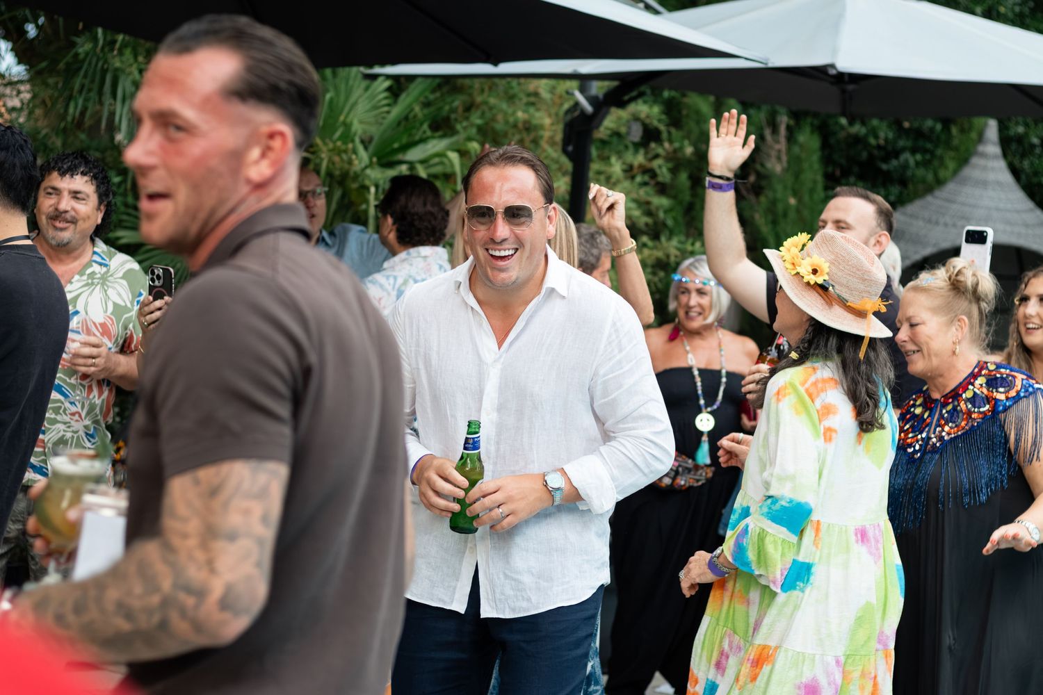 A smiling man in a white shirt holds a beer bottle while socializing at a lively outdoor party event.