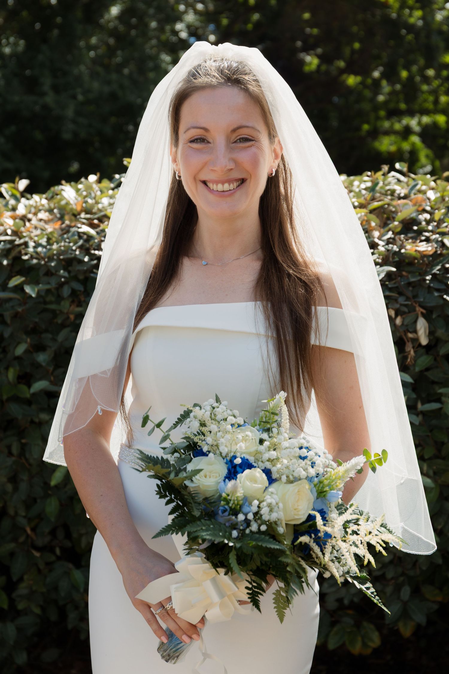 Smiling bride in white gown and veil holding blue and white floral bouquet outdoors near green hedges.