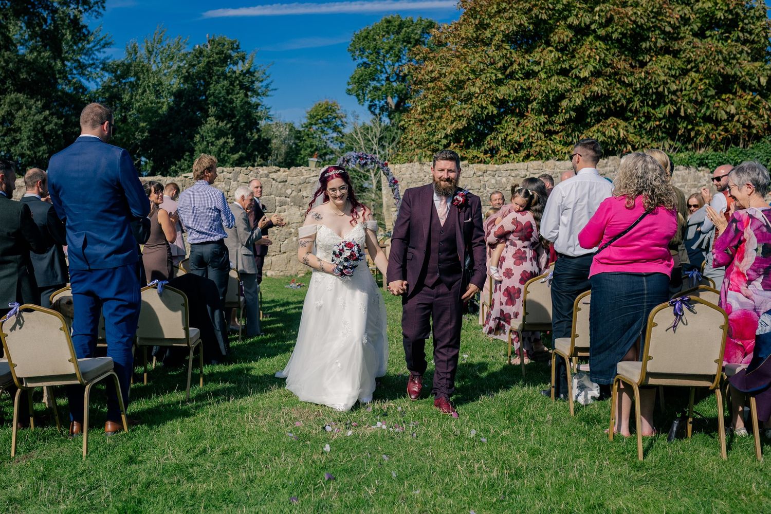 Bride and groom walk down outdoor wedding aisle surrounded by guests on a sunny day at a stone wall venue.