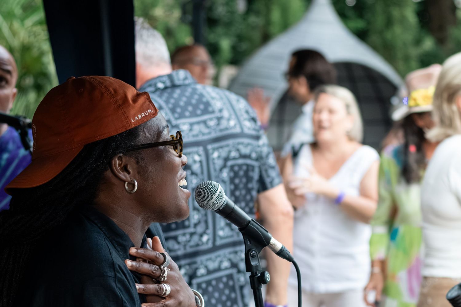A Black woman singer performs at an outdoor music festival, speaking into a microphone wearing an orange cap and sunglasses.