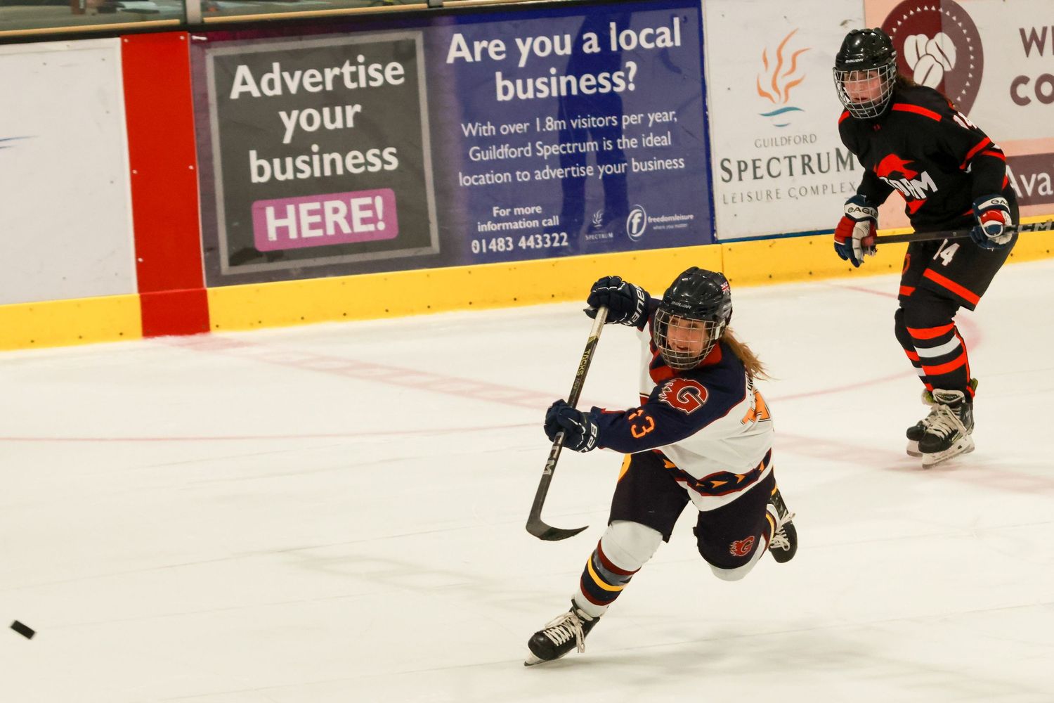 Young female ice hockey player takes a shot on ice rink with opponent skating behind her.