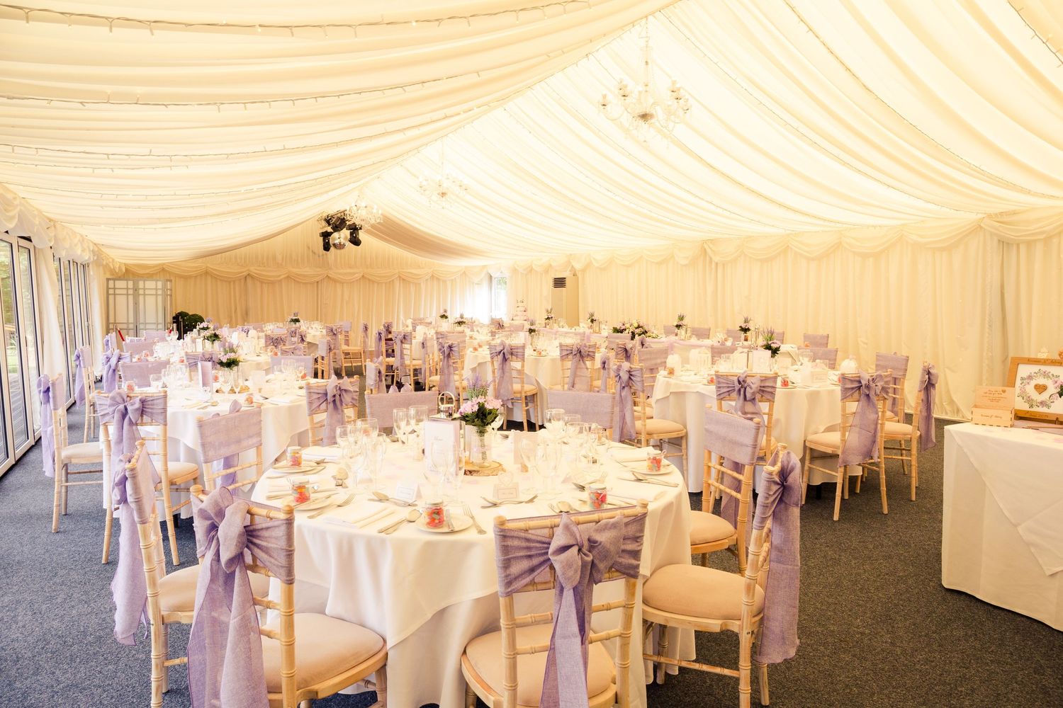 Elegant wedding marquee with lavender chair sashes, round tables, white linens, and draped cream ceiling decor.