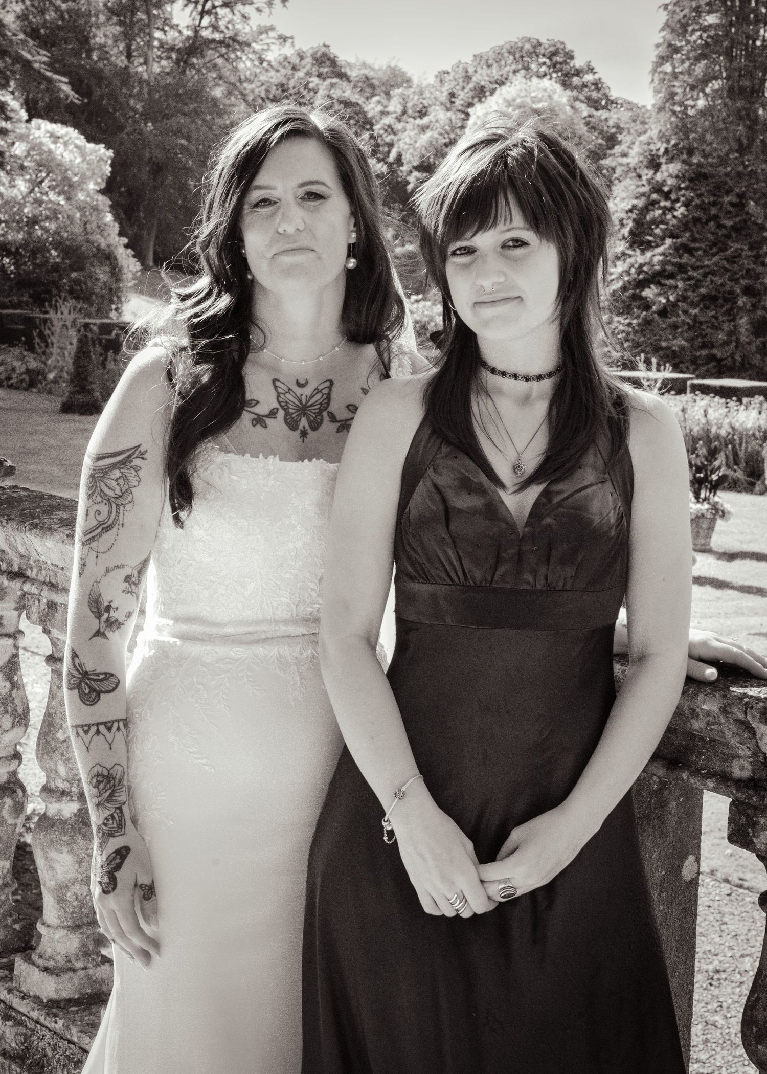 Black and white photo of two women posing together outdoors, one in a white wedding dress with tattoos, the other in a black dress.