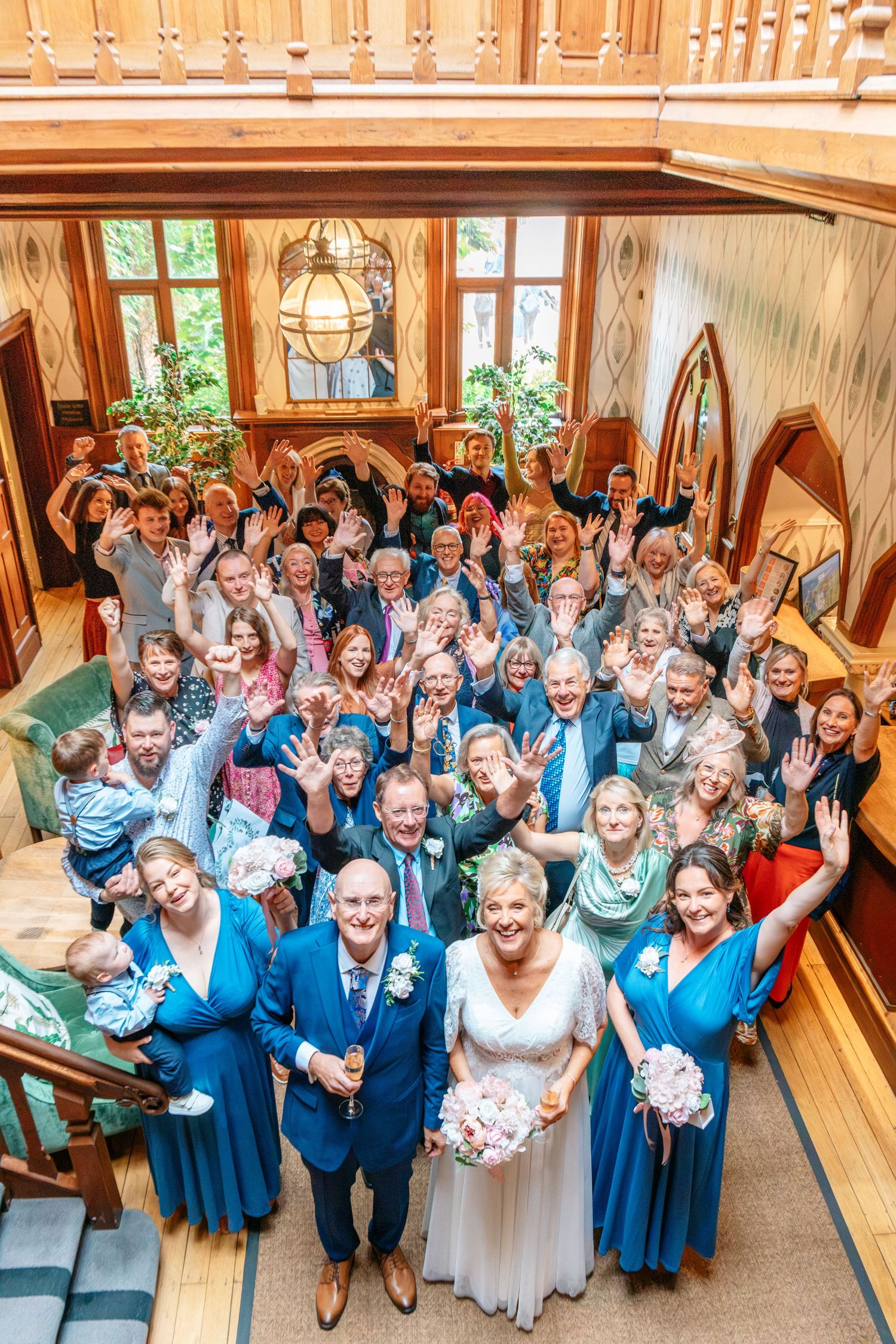 Wedding group photo with bride, groom, bridesmaids in blue, and guests waving in an elegant wood-paneled venue.