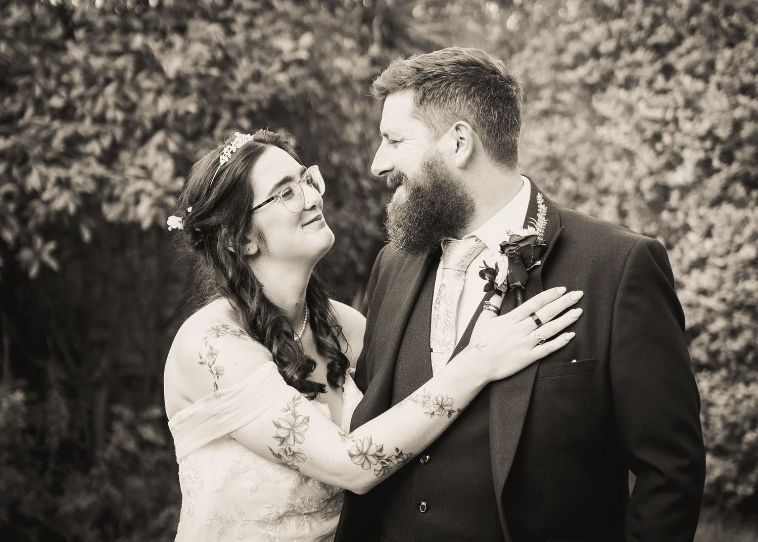 Black and white wedding photo of smiling bride with floral crown and tattooed groom with beard in suit.