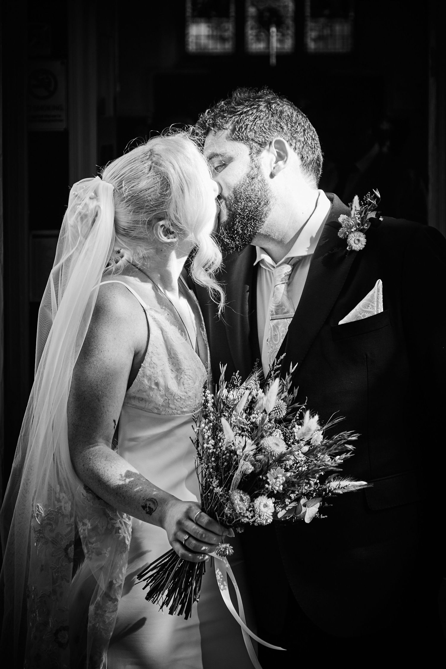 Black and white portrait of newlyweds kissing, bride in veil holding rustic bouquet, groom in formal suit.