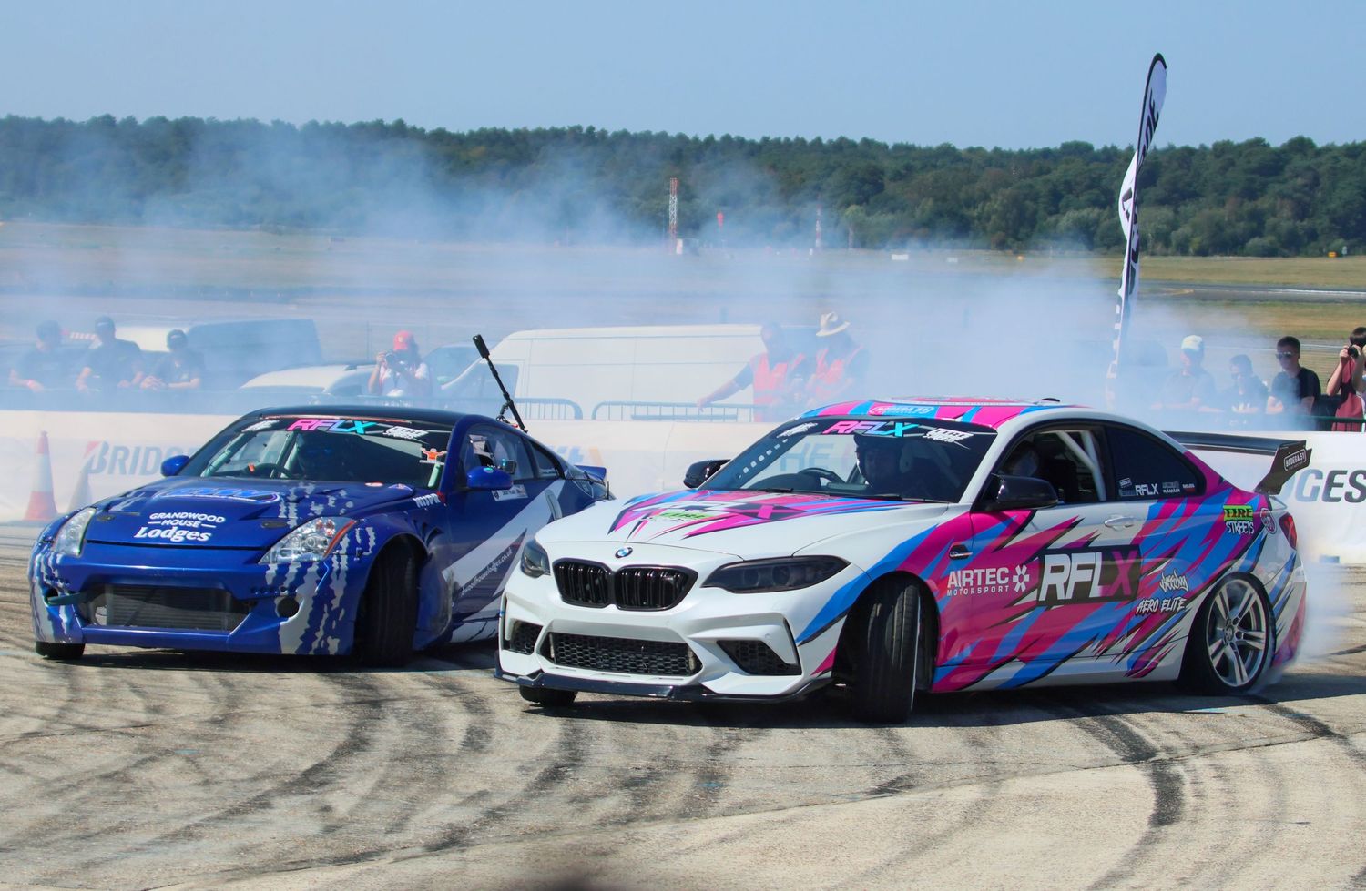 Two drift cars, a blue Nissan and pink BMW M2, battle through smoky turns at a motorsport event.