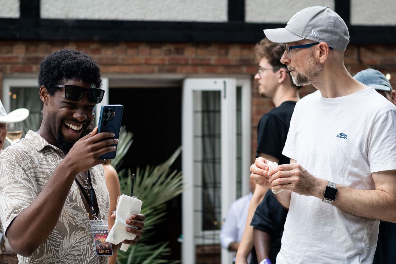 Two men laugh together outdoors, one holding a smartphone, at a casual daytime social gathering.