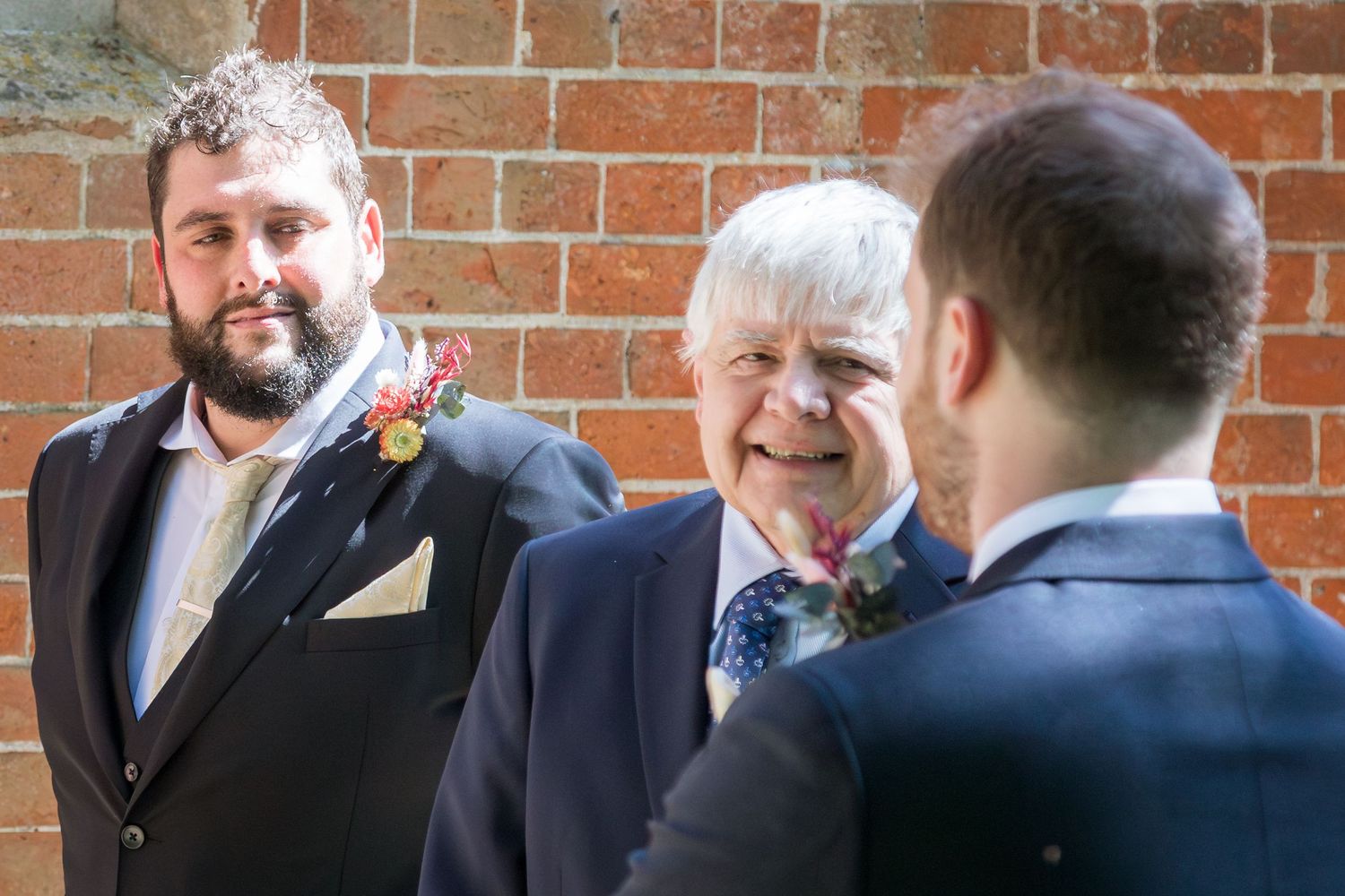 Bearded groom and older gentleman smiling together against brick wall at wedding.