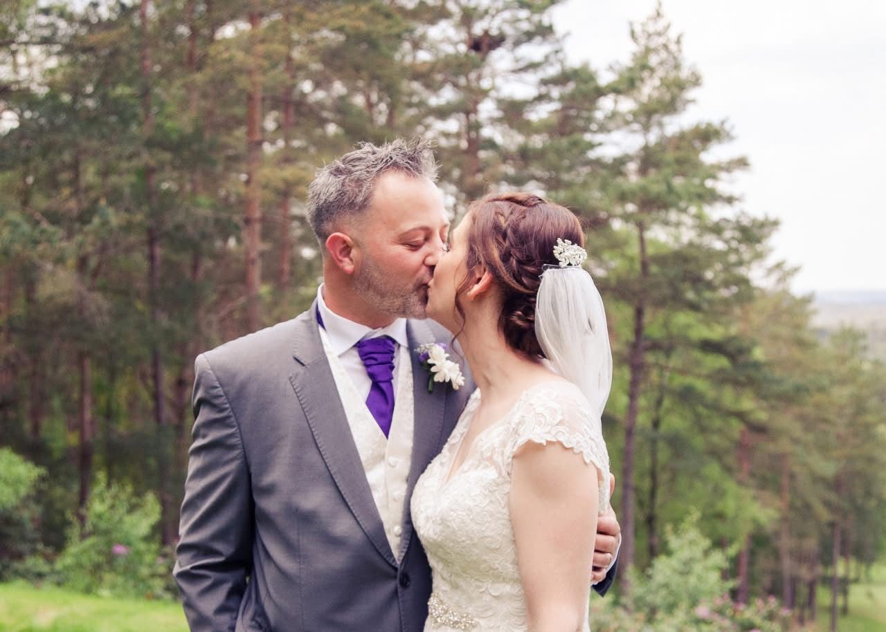 Bride and groom sharing tender kiss outdoors with pine forest backdrop on wedding day.
