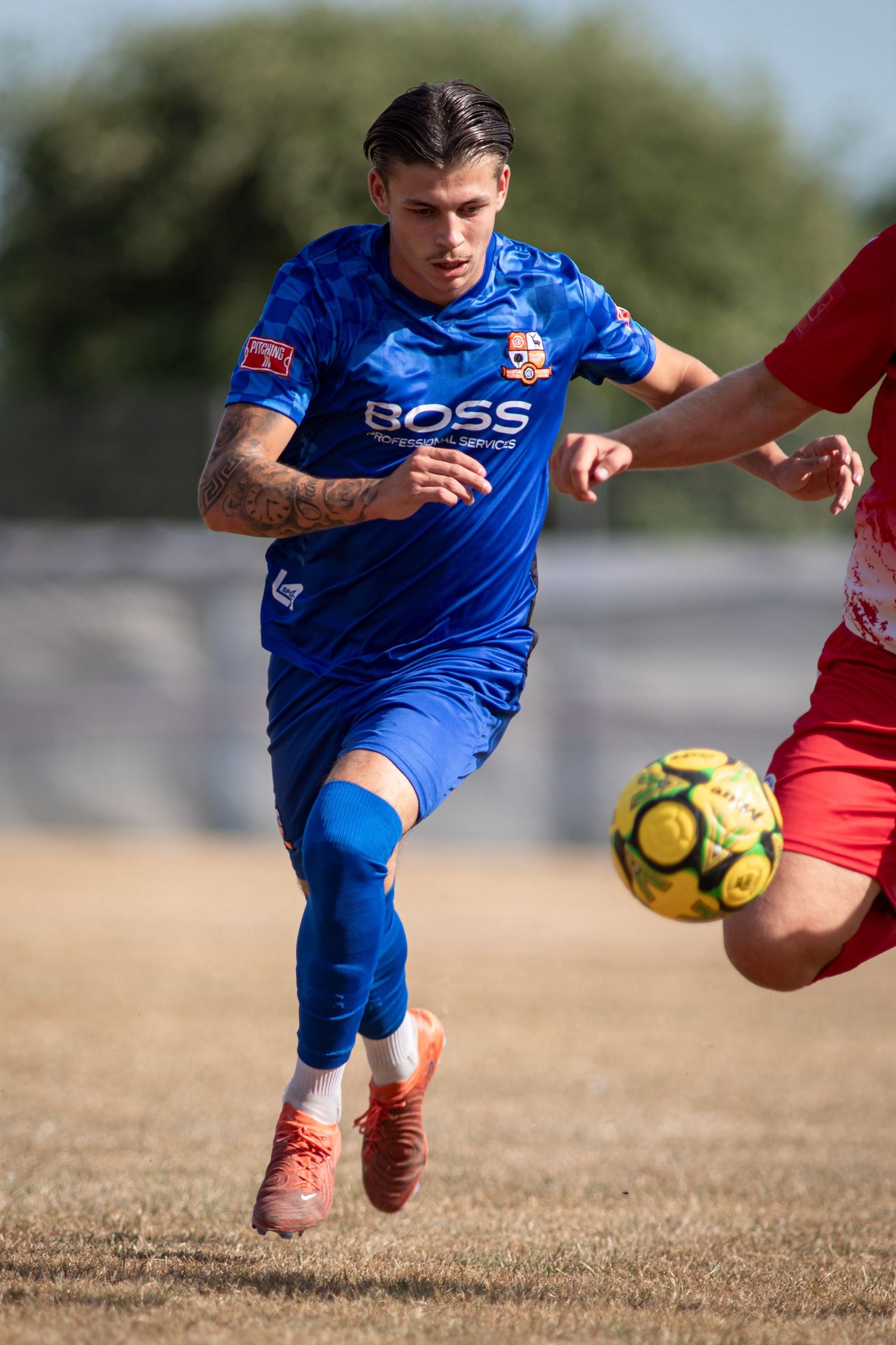 A footballer in a blue BOSS sponsored jersey chases the ball during a competitive outdoor match.