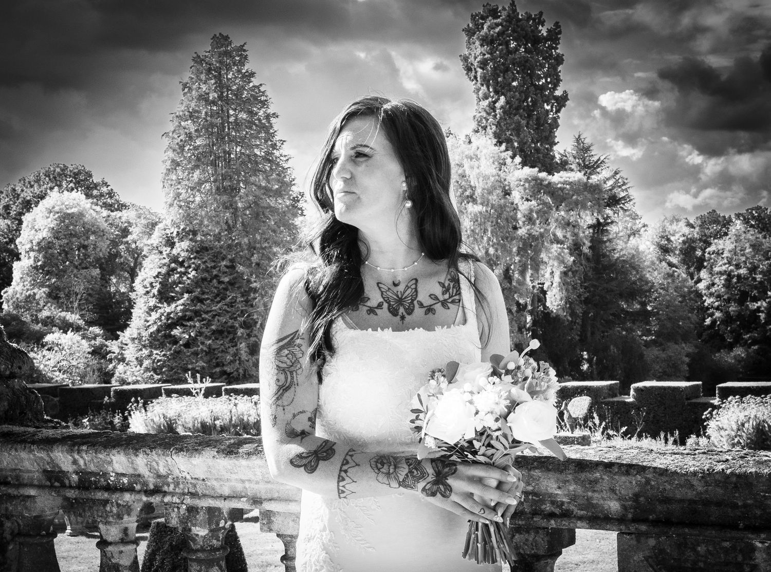 Black and white photo of a tattooed bride holding a bouquet in a garden with dramatic cloudy skies.