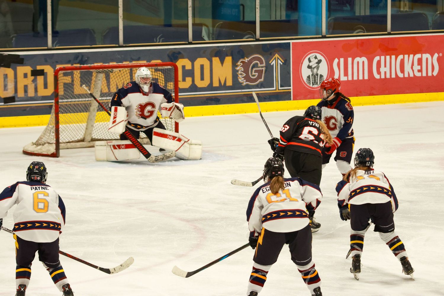 Women's ice hockey game action with goalie defending net while players compete near the blue line on indoor rink.