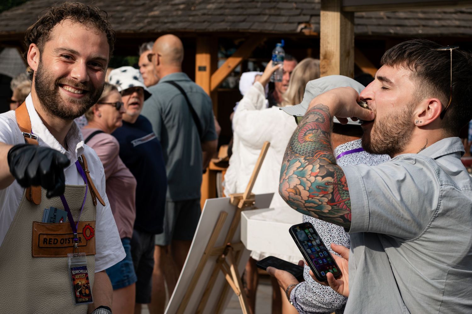 Two smiling men at an outdoor event, one wearing an apron stands by an easel while another with tattoos looks on.